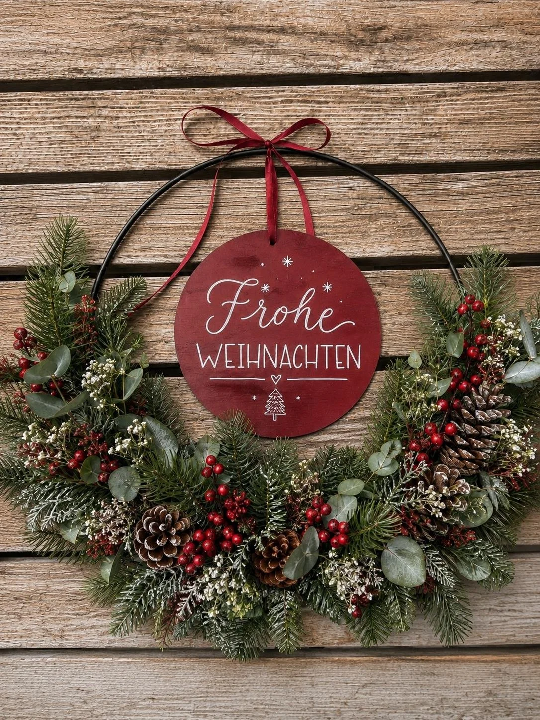 Weihnachtskranz aus Tannenzweigen, Eukalyptus, roten Beeren und Tannenzapfen in einem schwarzen Metallring vor einer Holzfassade mit rotem Anhänger Text auf Bild: Frohe Weihnachten