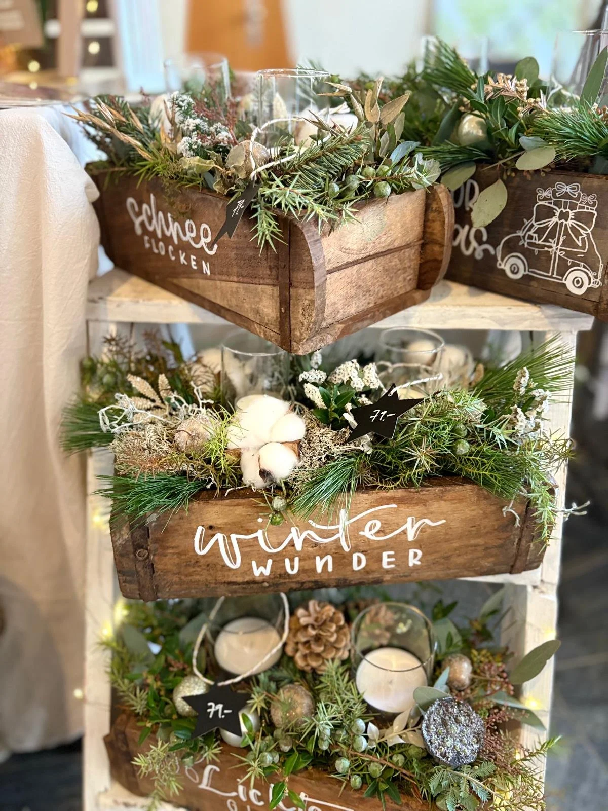 Drei rustikale Holzkisten mit Tannenzweigen, Kerzen, Christbaumkugeln, Baumwollzweigen und Preisetiketten in einer Weihnachtsdekoration Text auf Bild: Schöne Glöckchen, winter WUNDER, Fr.-