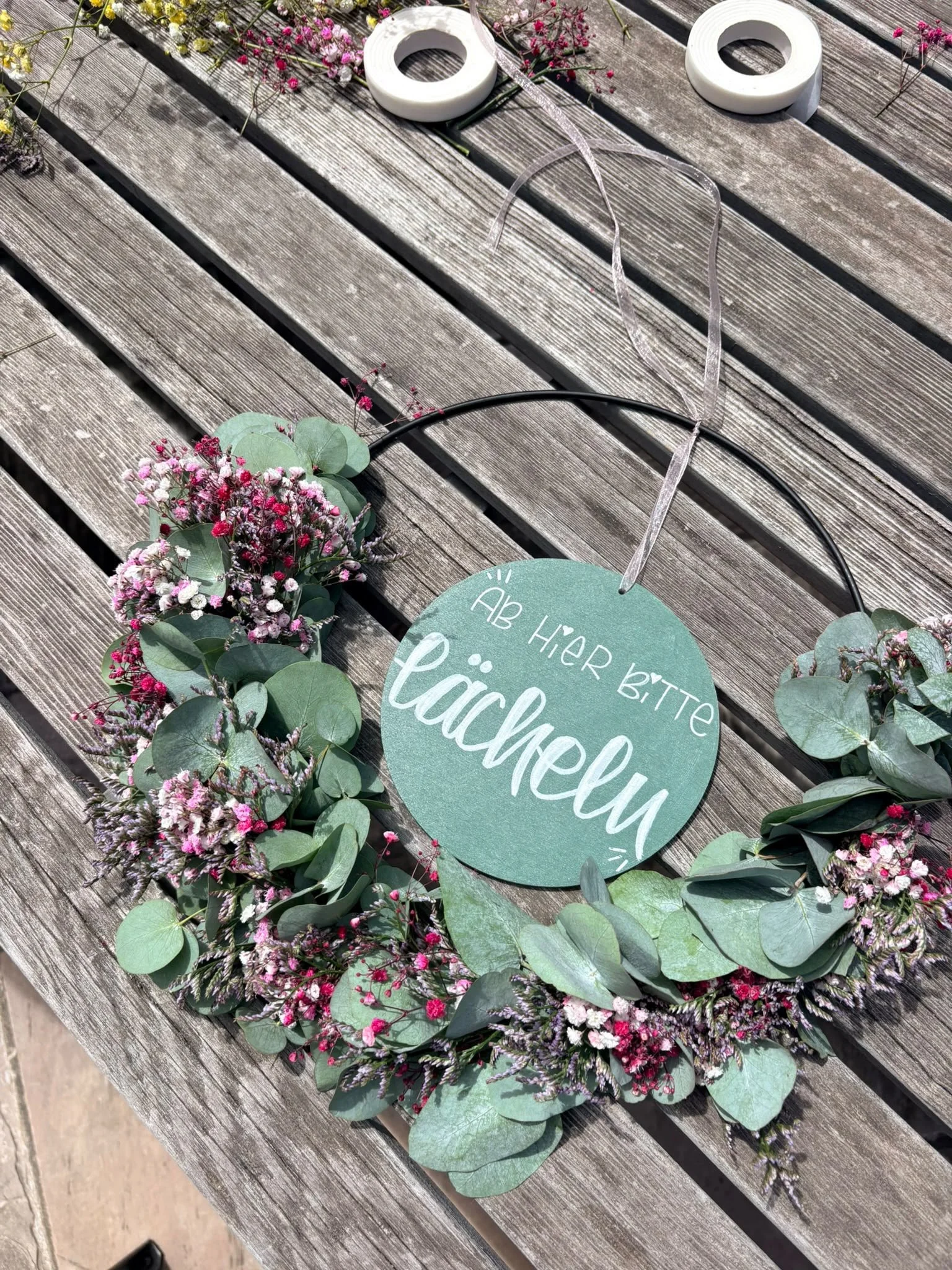 Rundes Schild mit Handlettering hängt an Trockenblumenkranz mit Eukalyptus auf Holztisch, Text auf Bild: Ab hier bitte lächeln