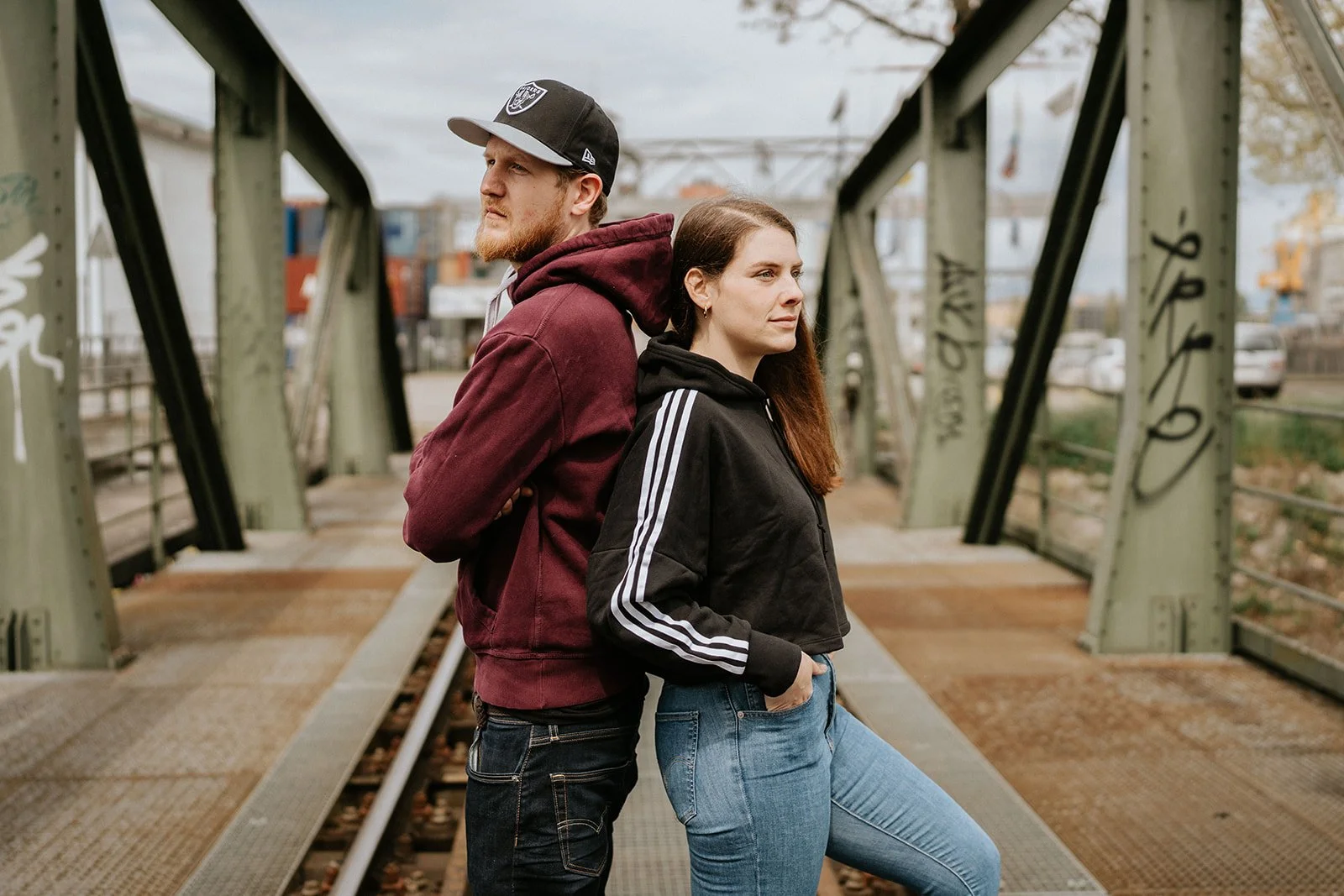 Zwei Personen lehnen Rücken an Rücken auf einer Fussgängerbrücke über Bahngleisen, eine mit bordeauxrotem Hoodie und schwarzer Kappe, die andere mit schwarzer Jacke mit weissen Streifen und Jeans