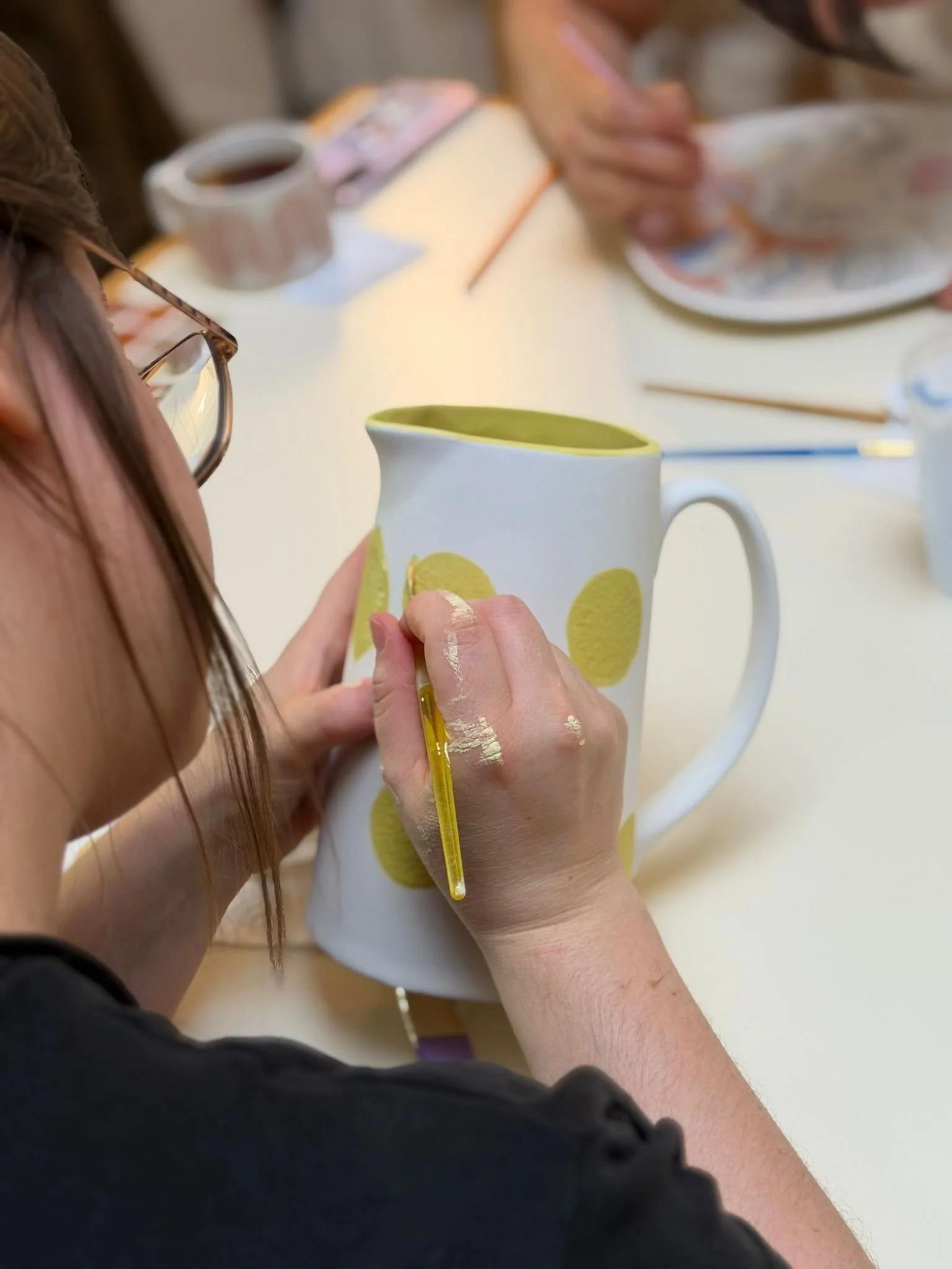 Person mit Brille bemalt einen weissen Krug mit gelben Punkten an einem Tisch, im Hintergrund gestaltet eine weitere Person einen Teller