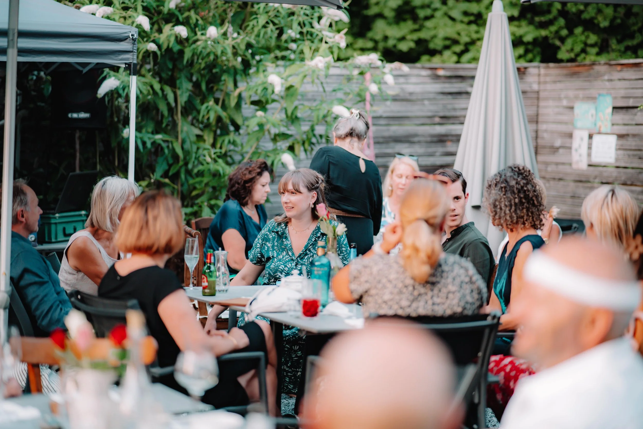 Gruppe von Personen sitzt und steht an Gartentischen im Freien, im Hintergrund blühende Sträucher und ein Sichtschutzzaun