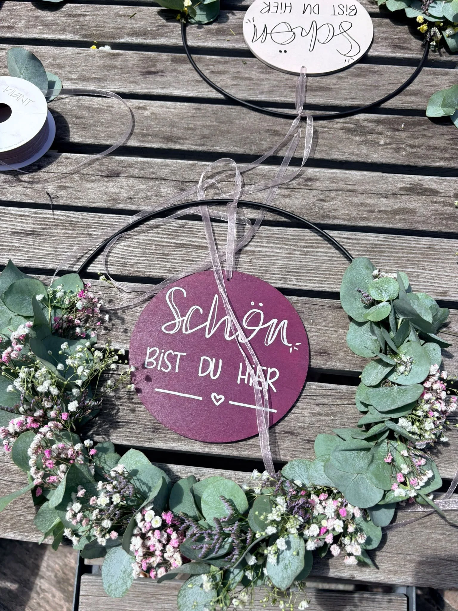 Zwei Trockenblumenkränze mit Eukalyptus und rundem Schild liegen auf Holztisch, im Hintergrund Bänder und weiteres Schild auf dem Kopf, Text auf Bild: Schön bist du hier