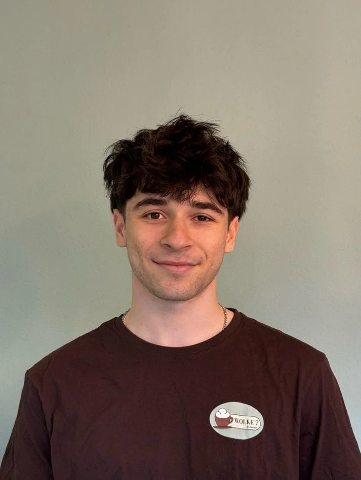 Person mit dunklem lockigem Haar und braunem T-Shirt vor hellgrüner Wand, Text auf T-Shirt: Wolke 7
