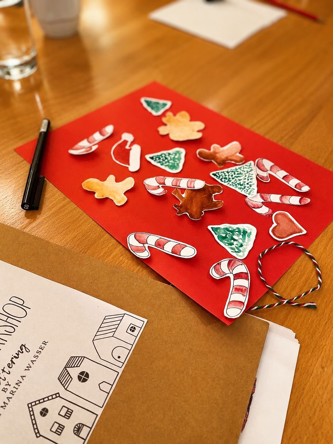 Rotes Papier mit ausgeschnittenen und bemalten Weihnachtsmotiven wie Zuckerstangen, Tannenbäumen, Sternen, Lebkuchenfiguren und Herz auf einem Holztisch neben Stift, Kordel und Workshop-Unterlagen