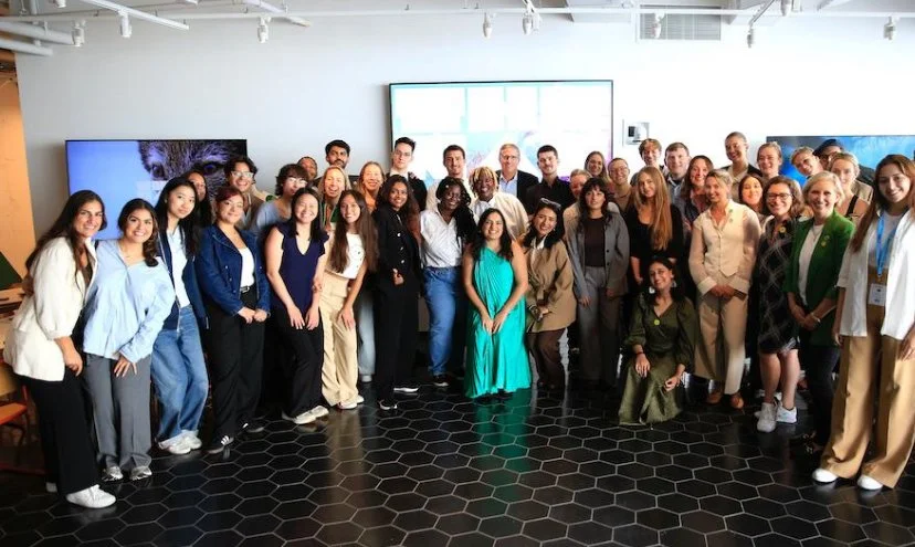 A large group of diverse people, mainly young adults, gathered in a modern room for a group photo. They are standing on black hexagonal tile flooring, with large screens on the wall behind them displaying a presentation or digital content. Some people are smiling and posing, indicating a professional or casual event or gathering.