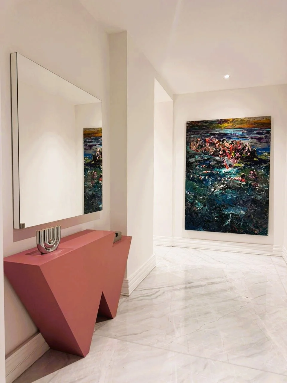 A modern art gallery with white walls and marble flooring. A large abstract painting with dark, bright, and colorful textured strokes hangs on the wall. A pink geometric console table holds a silver decorative piece and a tray, with a large mirror re