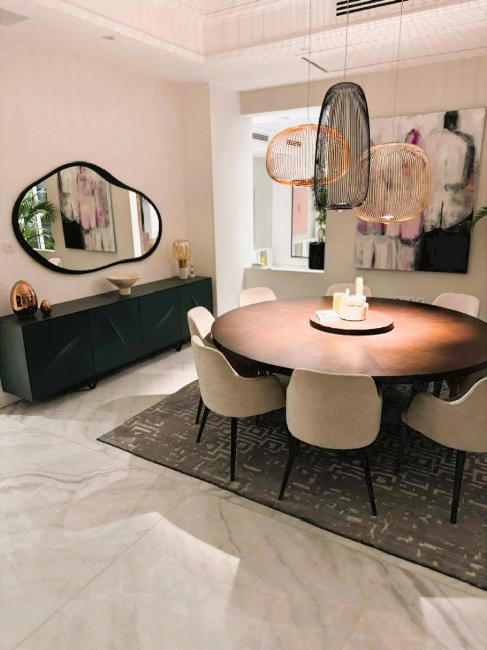 Modern dining room with a round wooden table, cream upholstered chairs, a black sideboard with a large mirror, abstract art, and hanging pendant lights.