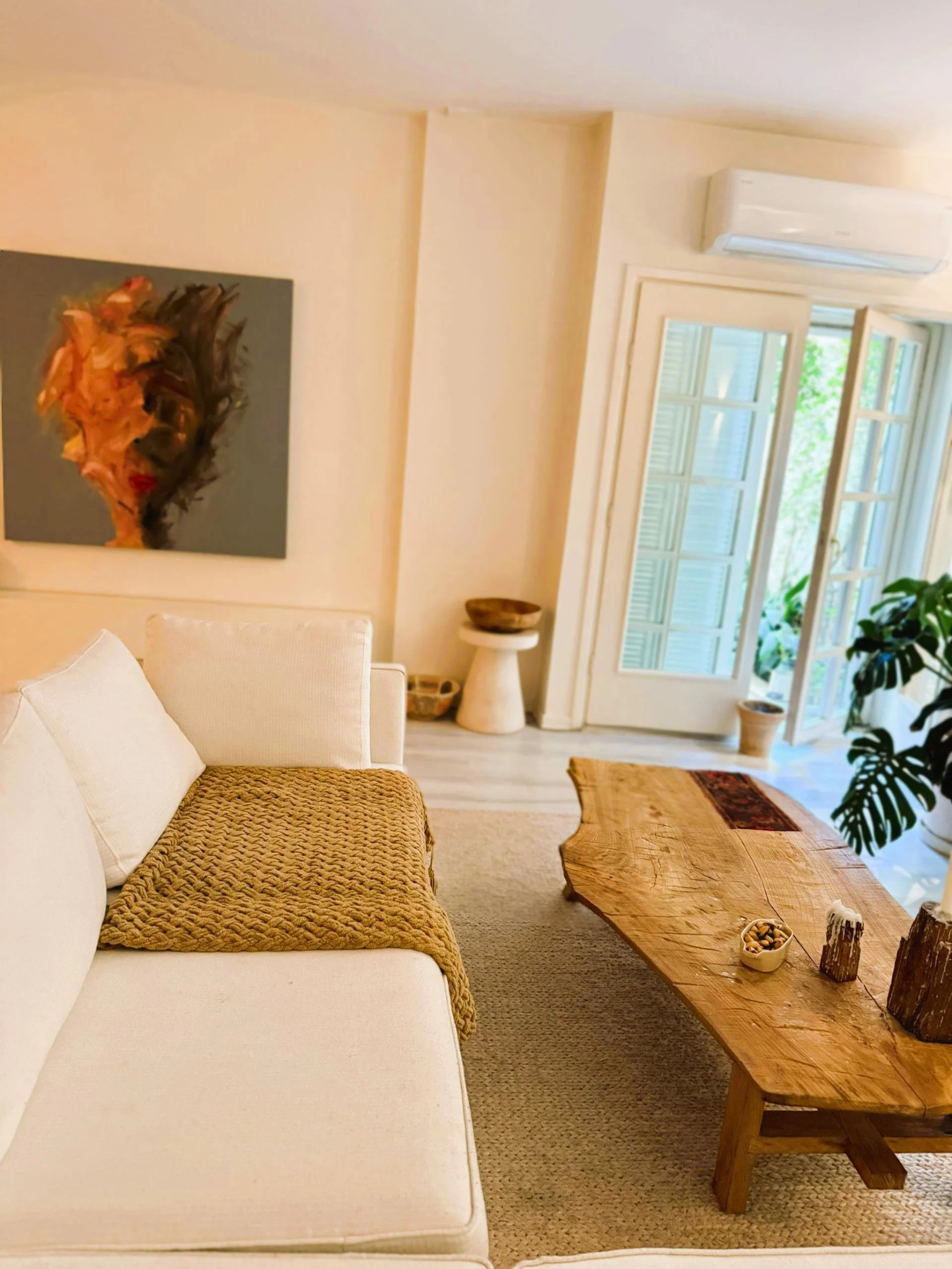 Living room with beige walls, a white sofa with a textured brown throw, a large rustic wooden coffee table, a colorful abstract portrait on the wall, and open French doors leading to a balcony with green plants.