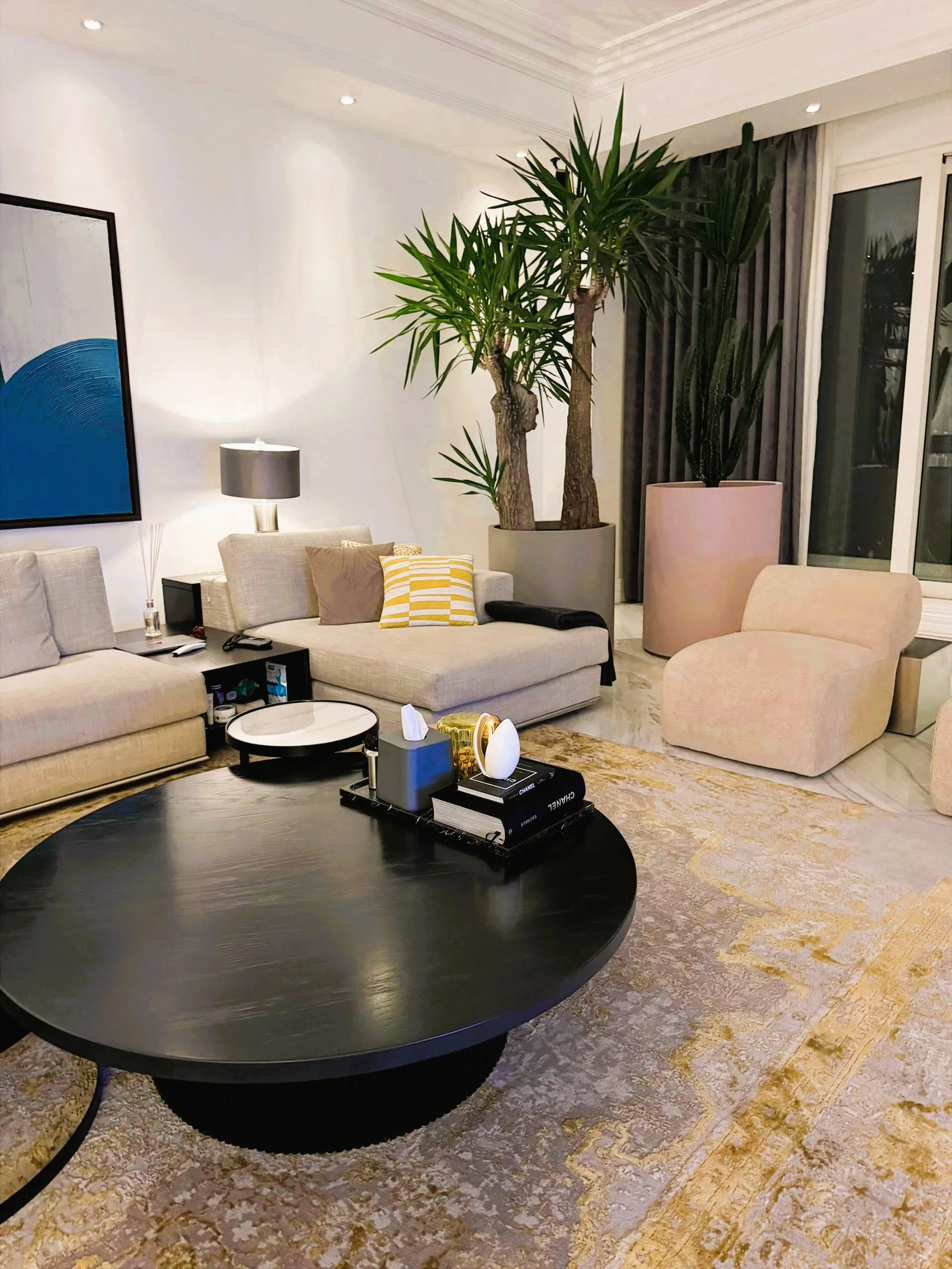Living room with beige sofas, a black coffee table, large potted plants, a floor lamp, curtains, and a colorful abstract painting.