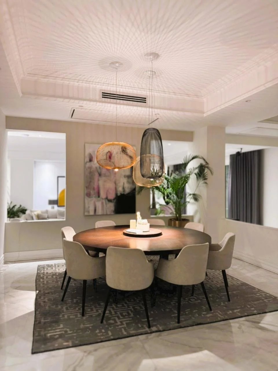 Modern dining room with a round wooden table, beige upholstered chairs, hanging woven pendant lights, a decorative rug, and indoor plants.