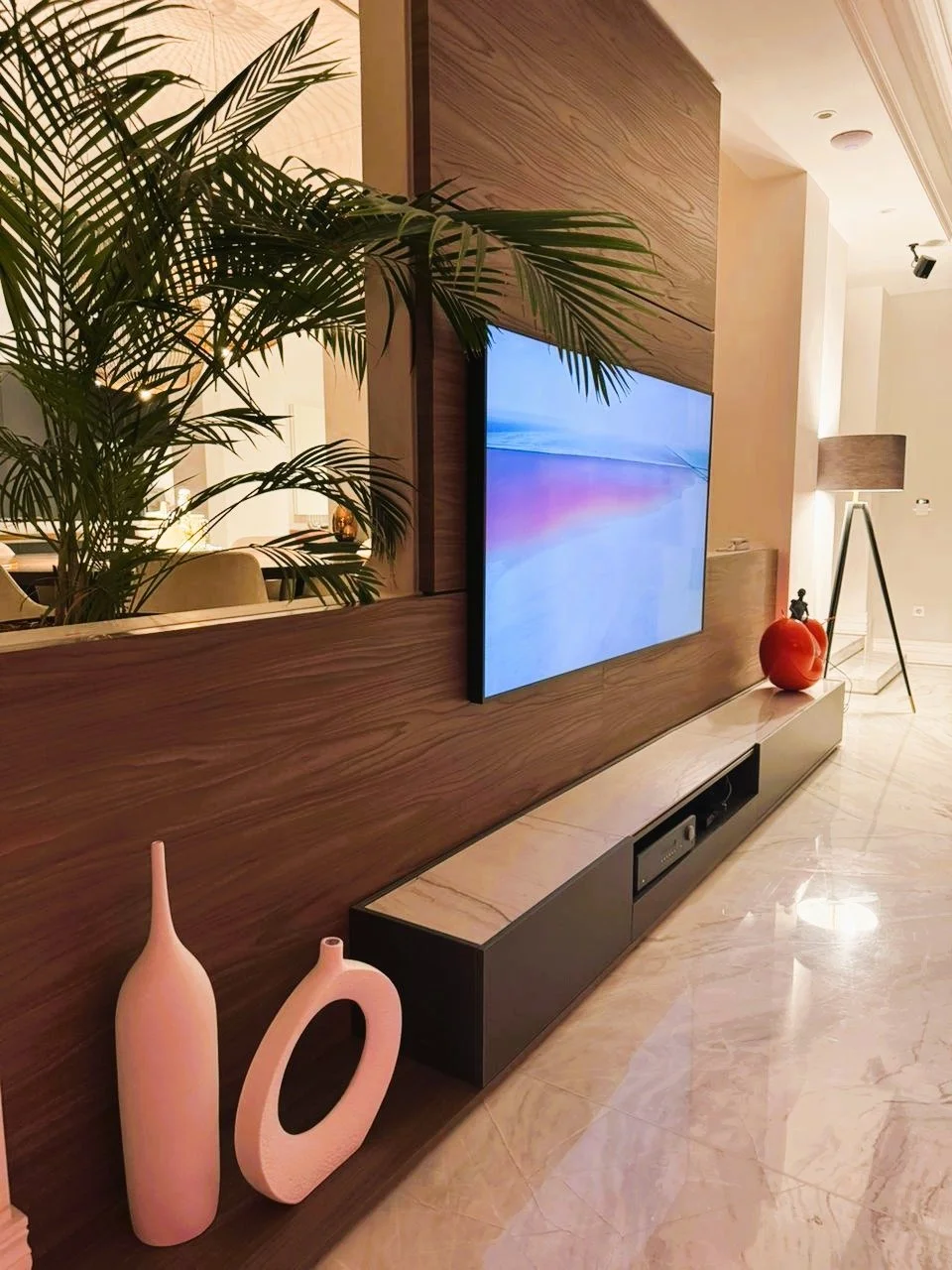 Living room with a flat-screen TV mounted on a wooden wall, decorated with pink vases, a red object, a small sculpture, tall floor lamp, and large green plant.