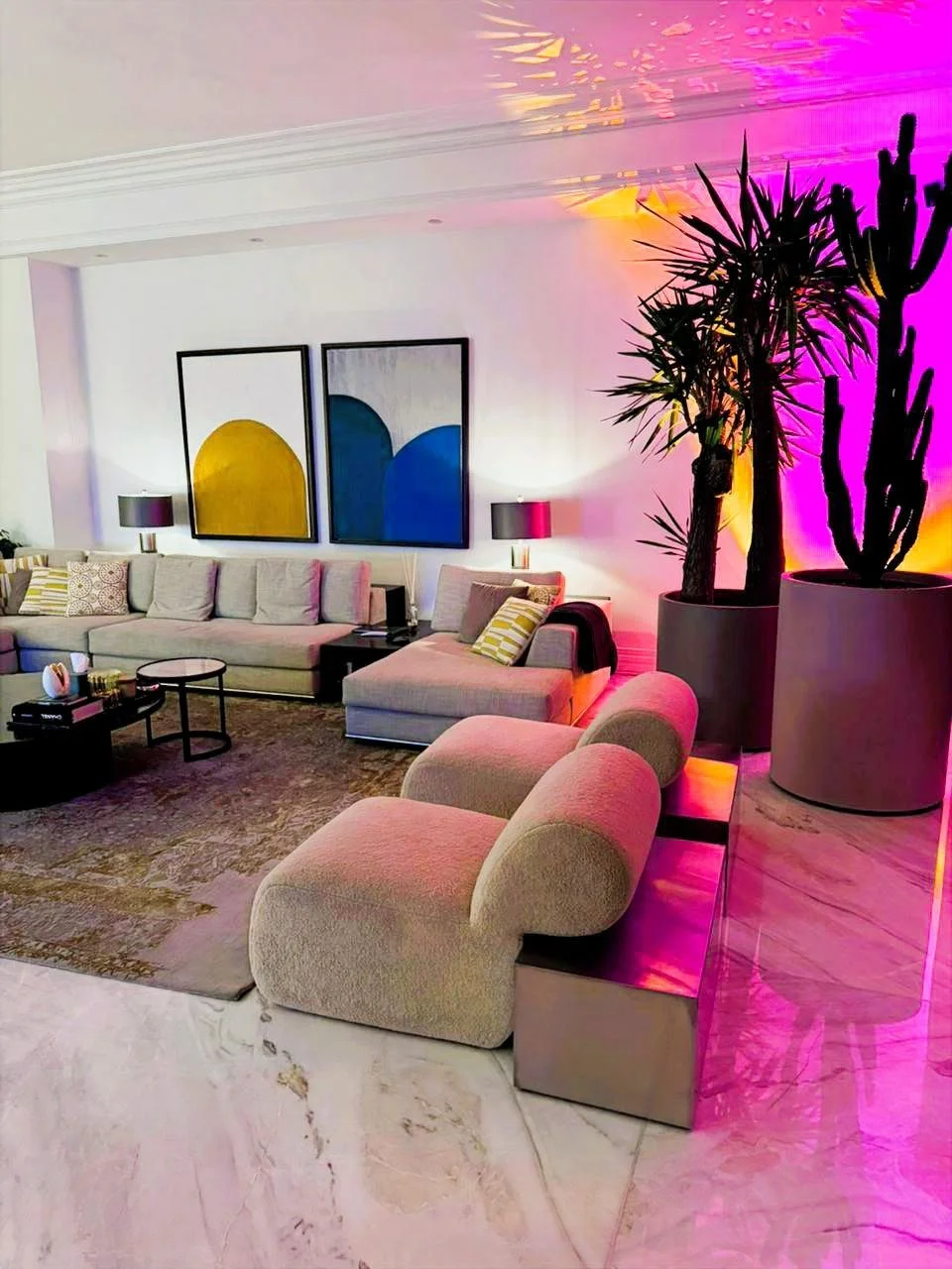 Modern living room with beige sectional sofa, two armchairs, a brown rug, and artwork with yellow and blue shapes on the wall. Large potted plants with tall, spiky leaves are illuminated by colorful pink, purple, and yellow lighting.