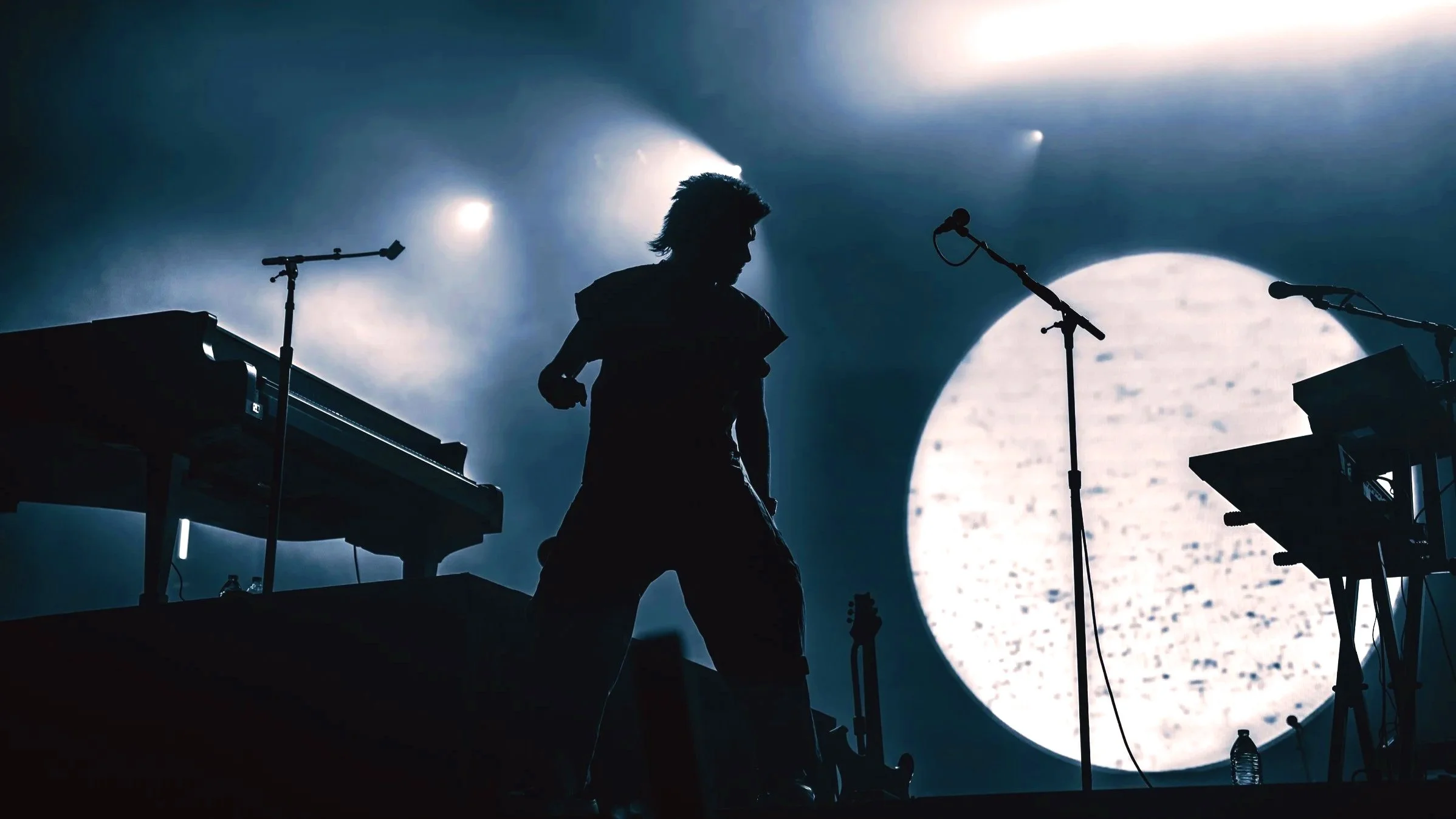 Musician performing on stage with keyboard and microphones silhouetted against a large circular screen or moon with bright lights.