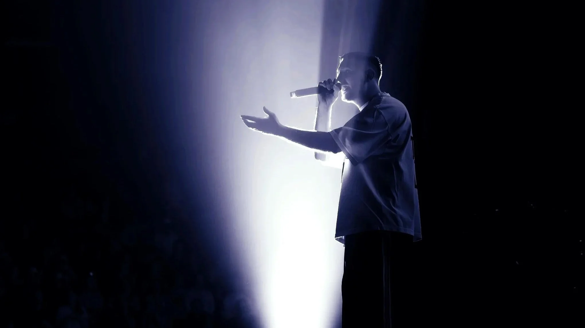 A man with short hair, wearing a dark shirt, singing or speaking into a microphone on stage, backlit by bright white light, creating a silhouette effect.