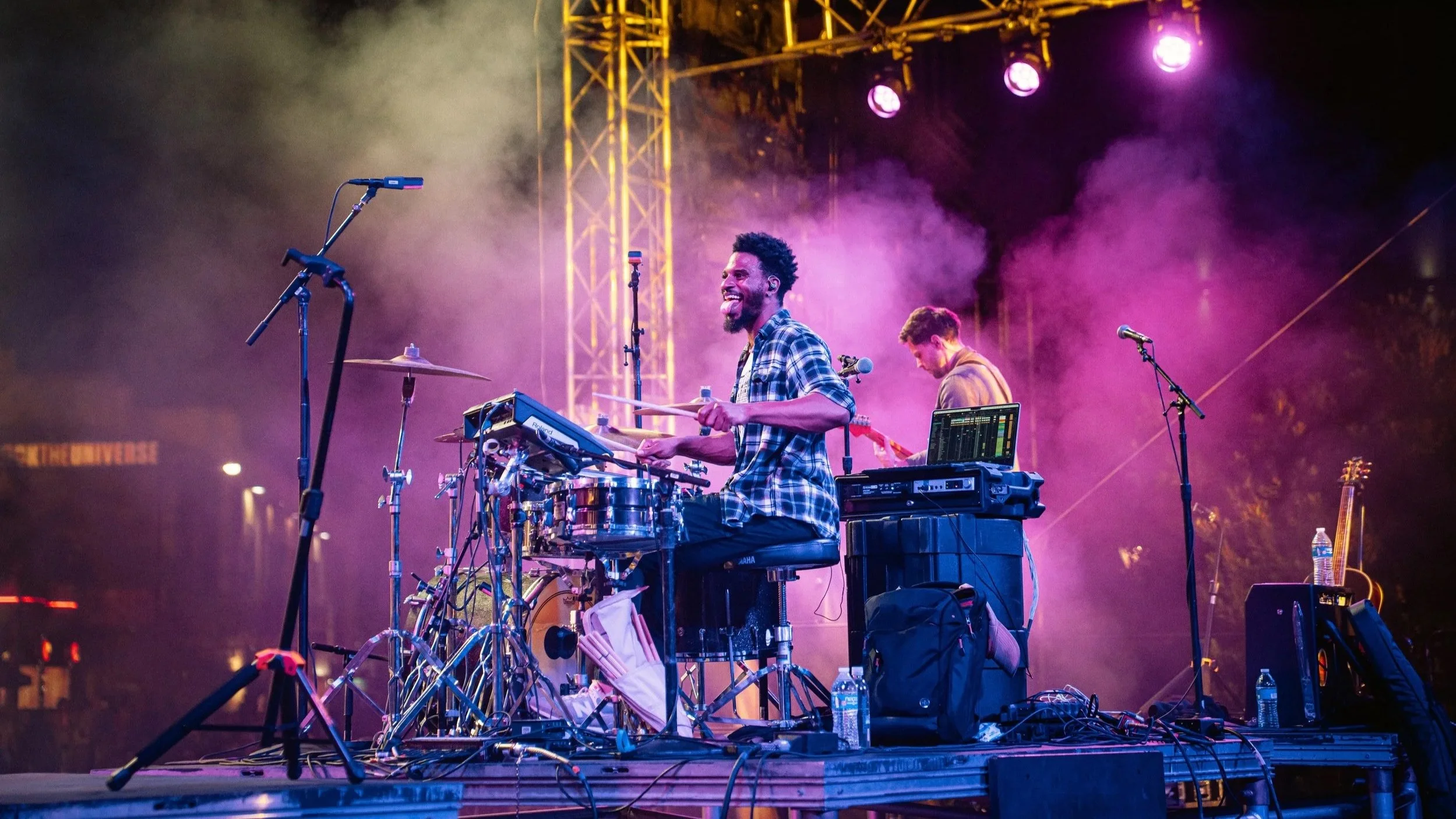 A musician playing a drum set on stage at night, smiling and enjoying the performance. The stage is lit with purple and yellow lights, with equipment like microphones and laptops around him.