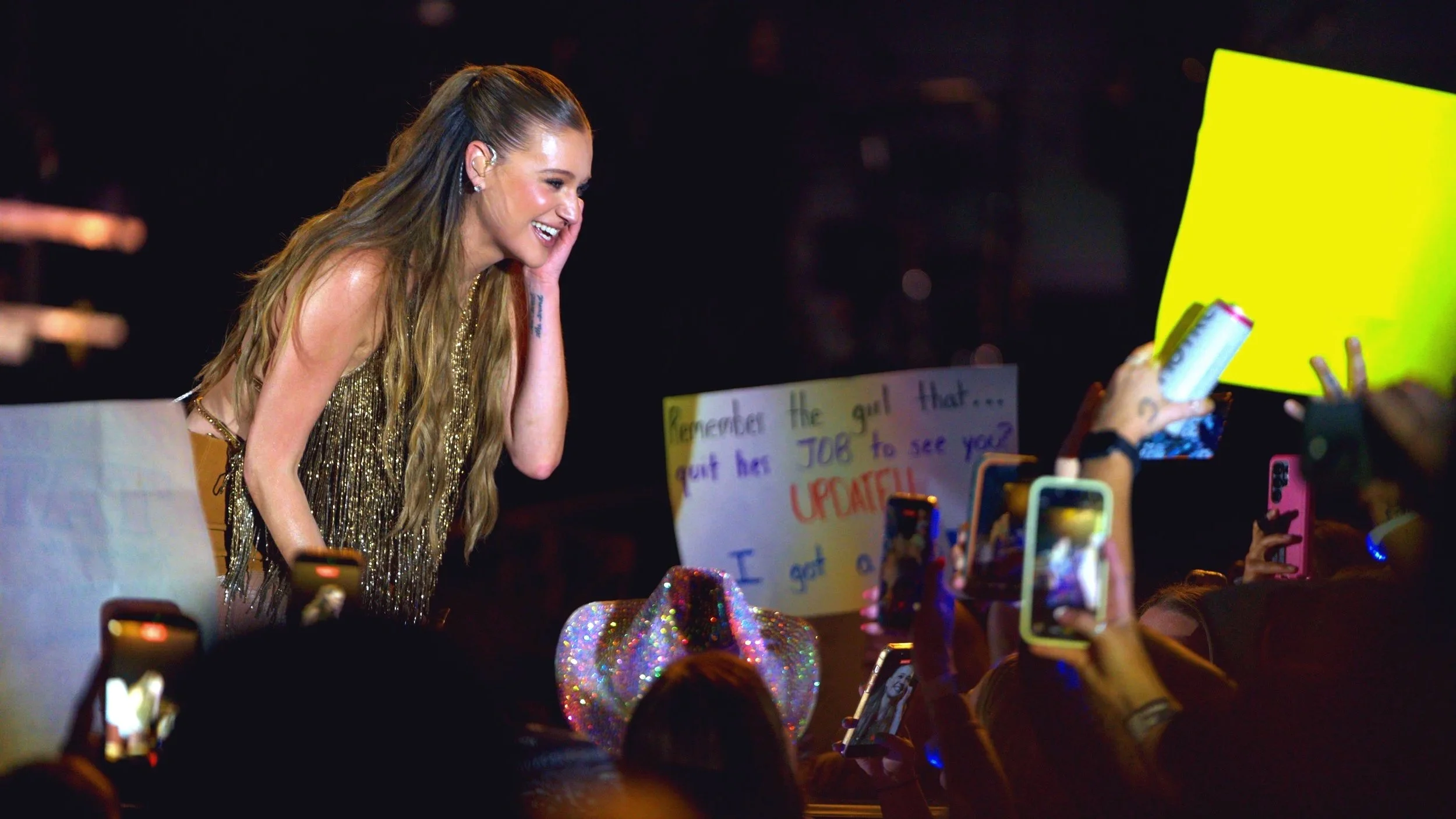 A woman with long wavy hair in a glittering dress smiling and leaning toward fans holding signs and taking photos at a concert.
