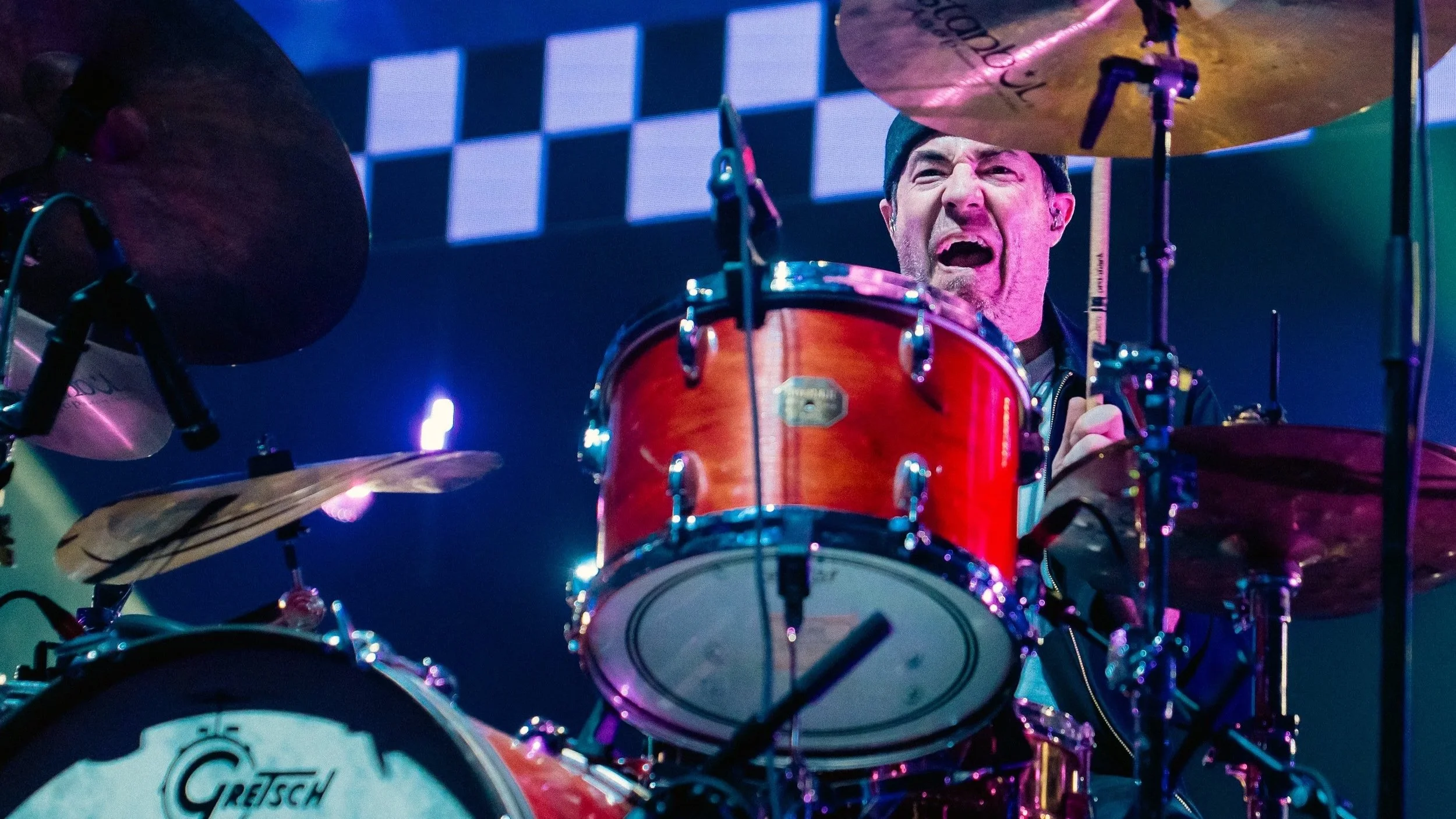 A man playing a red drum set on stage, with colorful stage lighting and a checkered background.