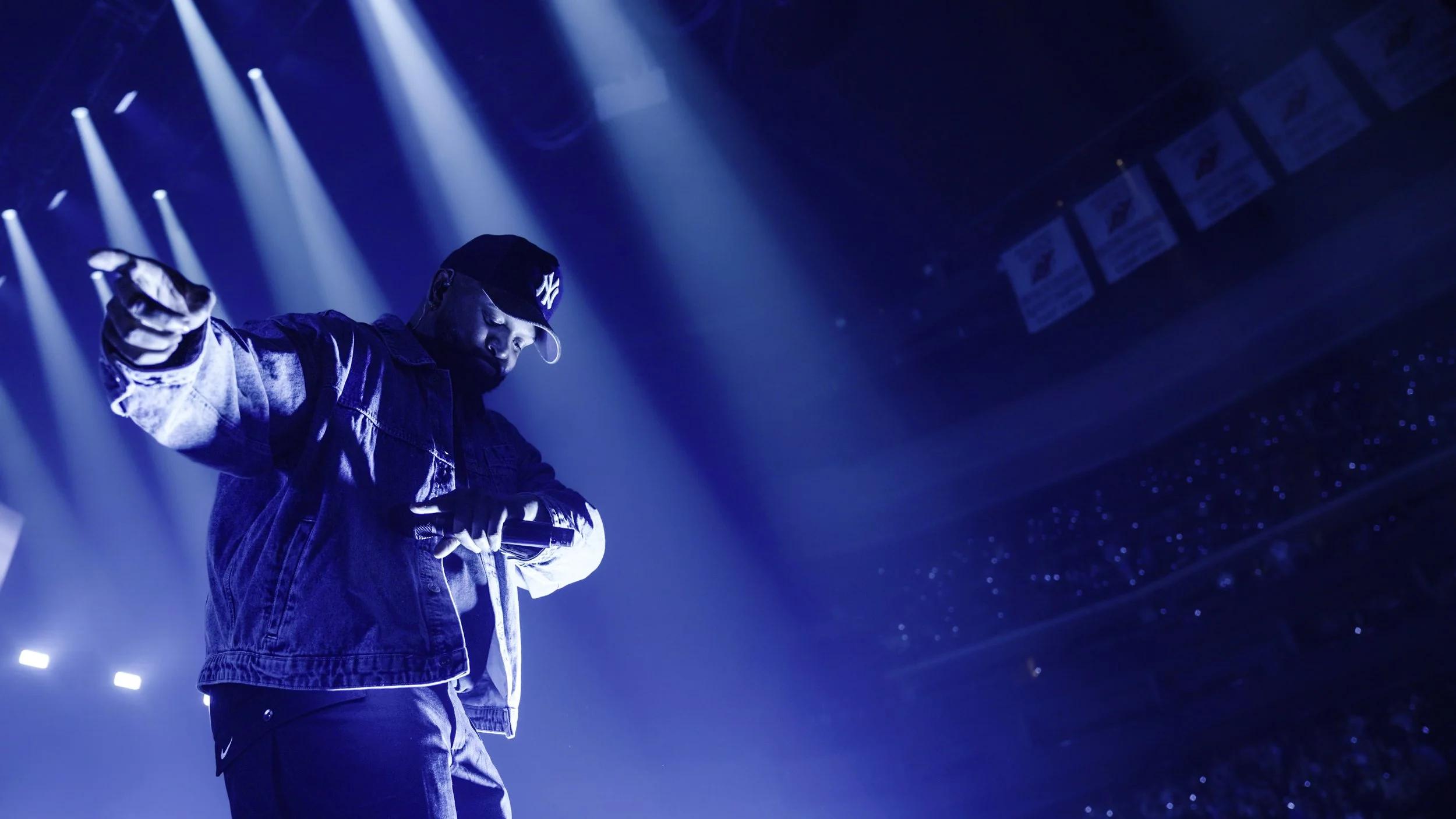 A male performer wearing a black cap and denim jacket on stage, illuminated by blue stage lights, holding a microphone.