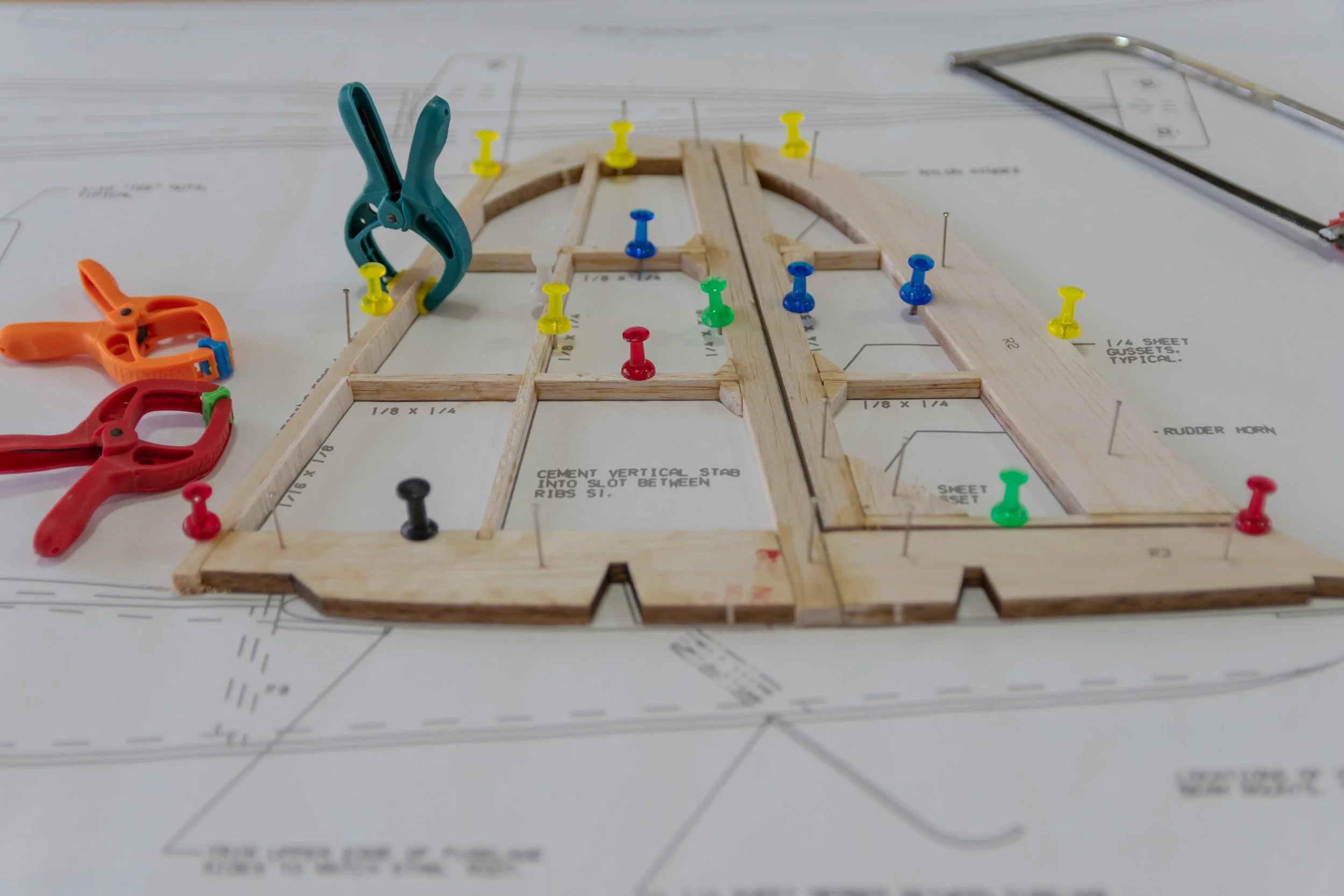 Wooden model aircraft wing with colored pins on top of construction blueprint and clamps on the side.