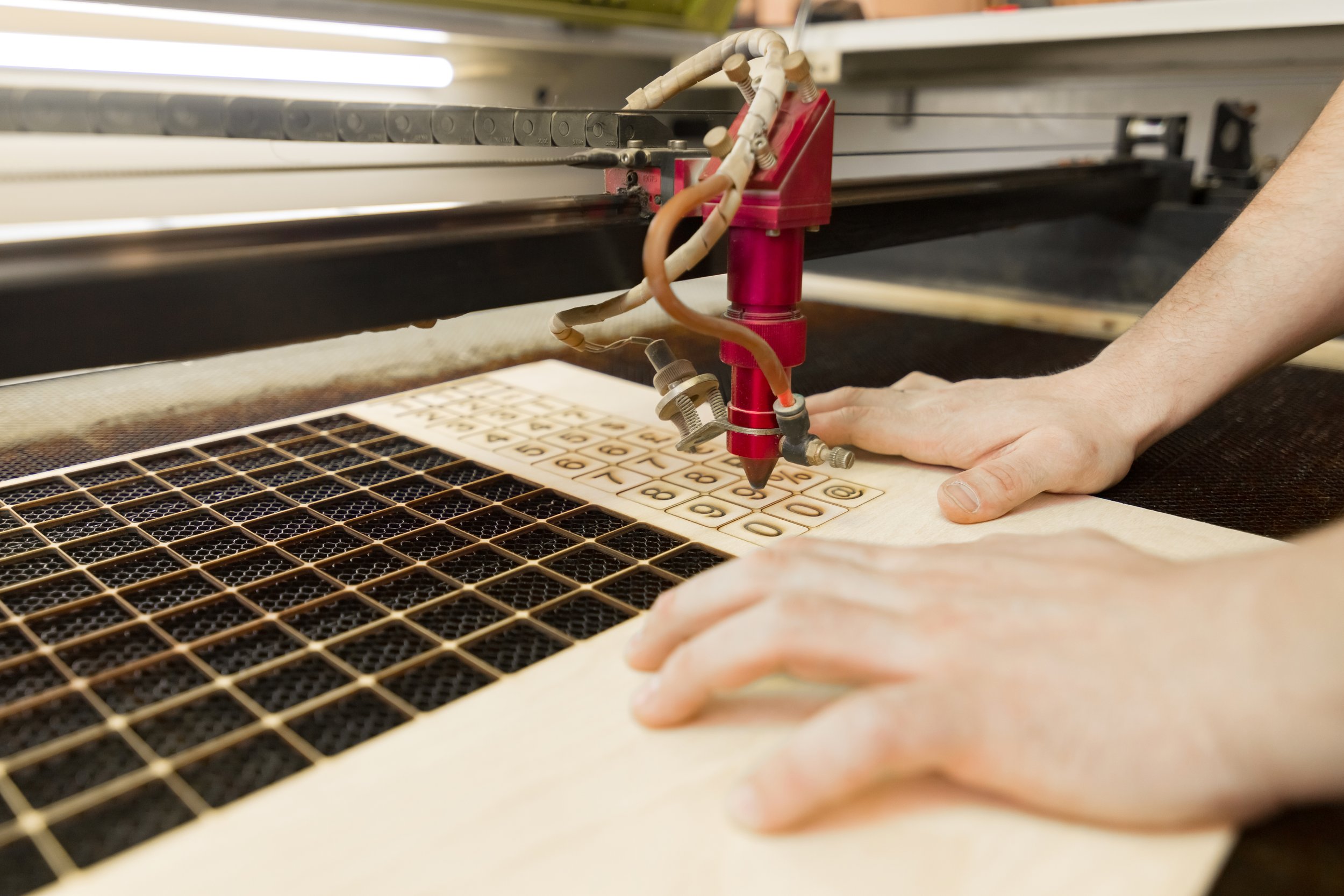 A laser cutting machine engraving numbers and symbols on a piece of wood. A person's hands are guiding the wood under the laser head.