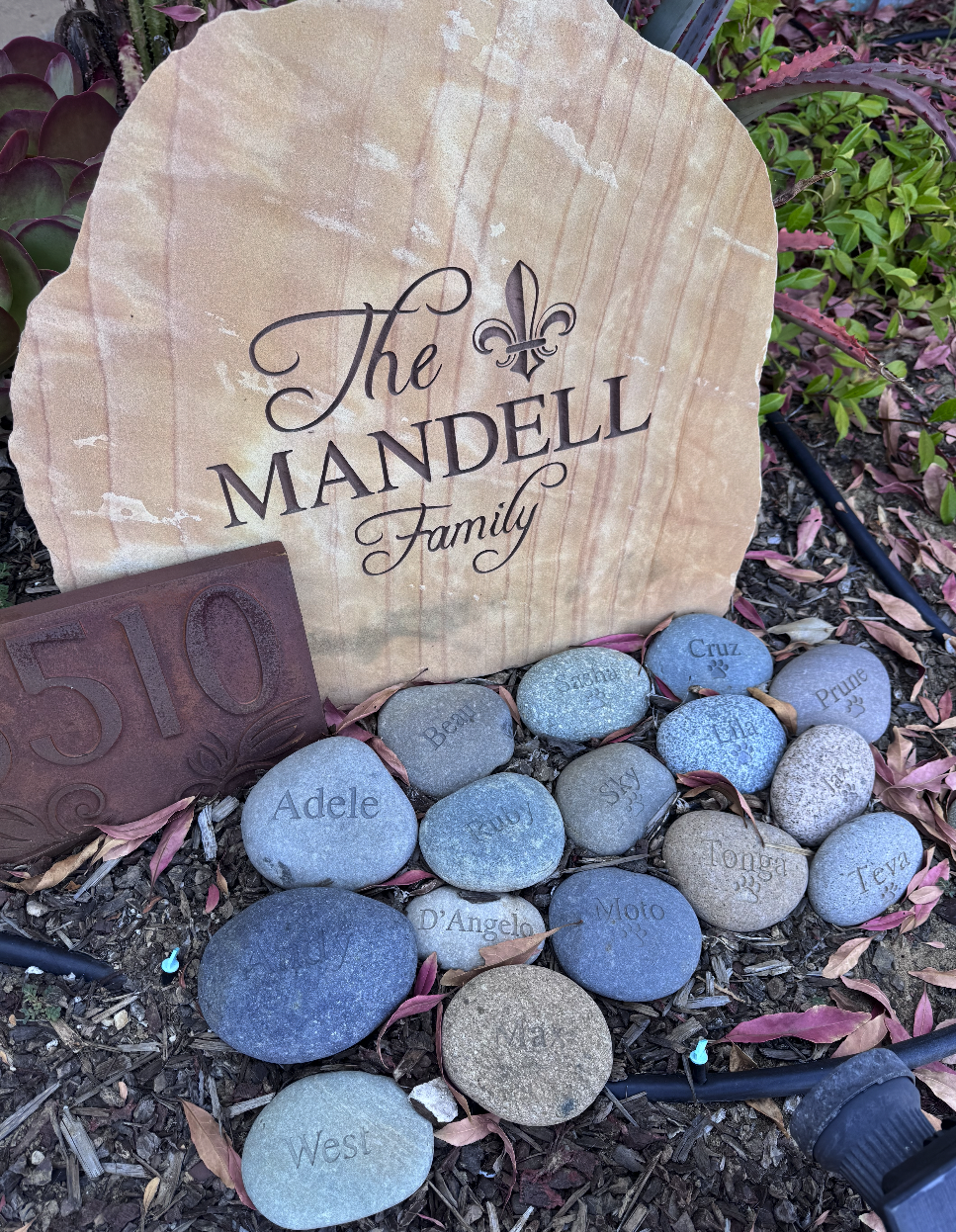 A garden decoration with a stone plaque reading 'Family', surrounded by engraved rocks with various names, a decorative metal engraved sign, and a small black electric watering device in the soil.
