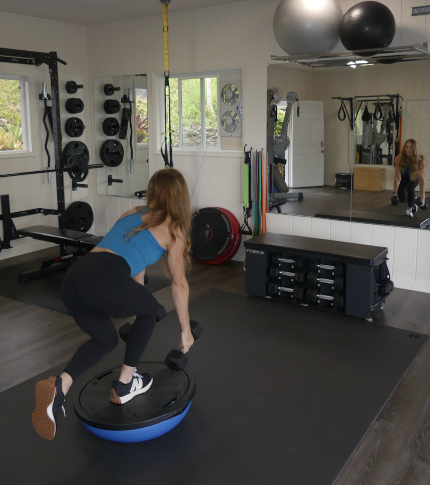 A woman in a gym using a balance trainer for exercise. The gym has various workout equipment including weights, resistance bands, exercise balls, and a large mirror.