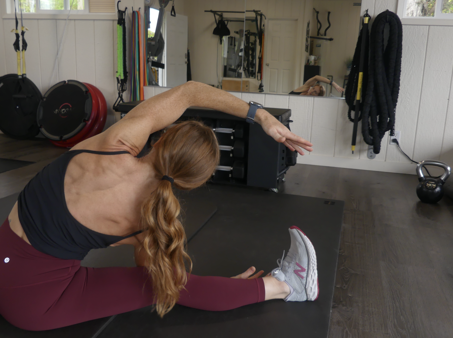 Woman stretching on a gym mat, wearing a black sports bra, maroon leggings, and New Balance sneakers in a private fitness studio with various exercise equipment in the background.