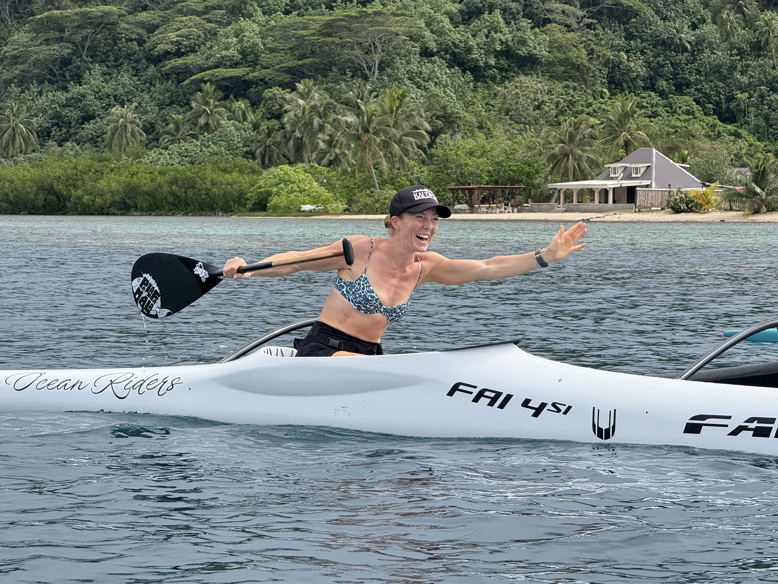 Woman in a sports bra and cap paddling an outrigger canoe on an ocean with lush green trees and houses in the background.