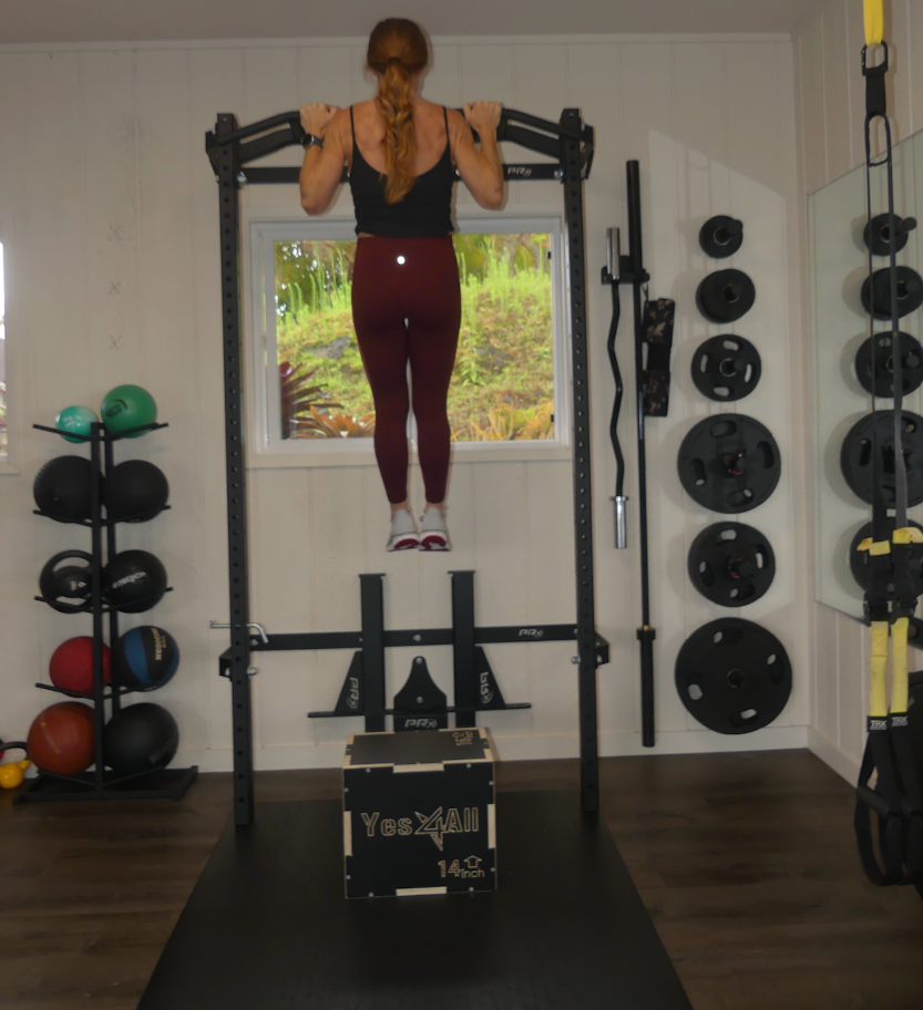 A woman doing a pull-up in a private studio space, hanging from a pull-up bar on a black power rack next to a window.