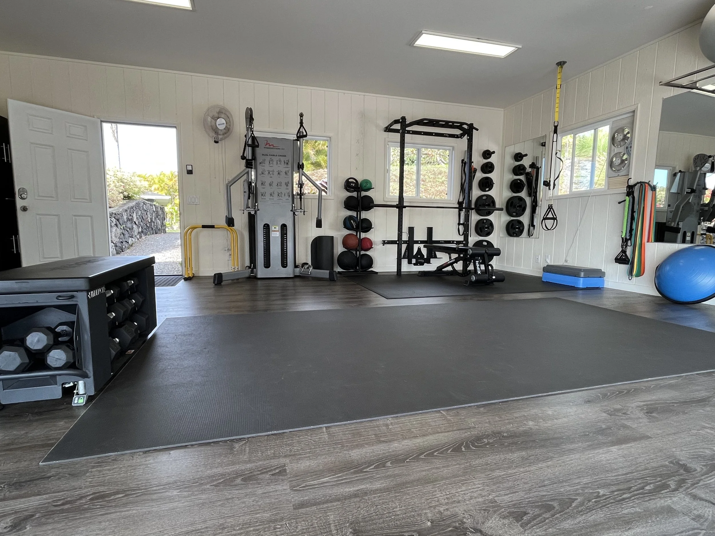 Inside a private fitness studio with weightlifting equipment, exercise balls, dumbbells, resistance bands, and a large black mat on the wooden floor.
