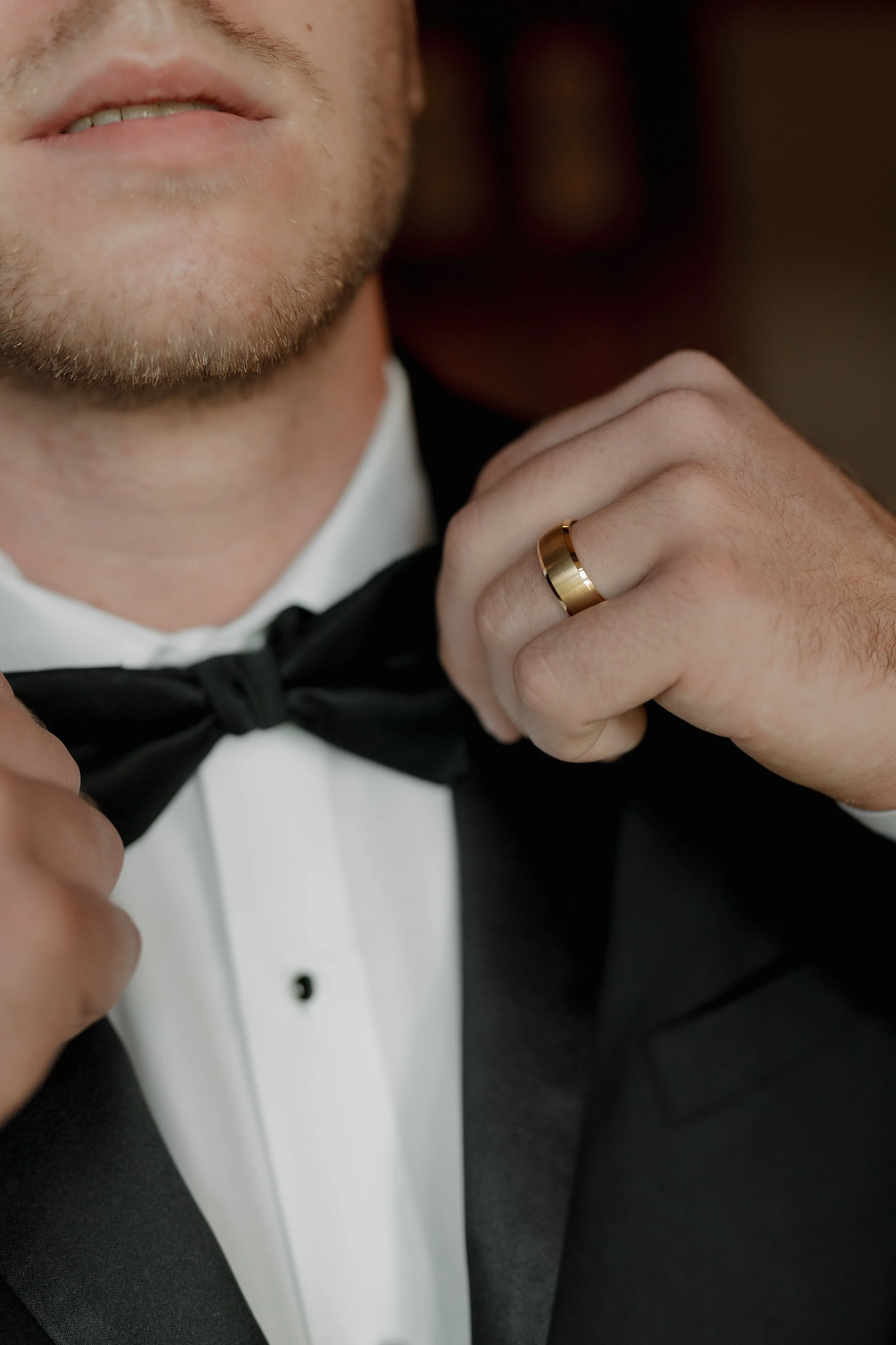 Close-up of a man in a tuxedo adjusting his black bow tie, with a gold wedding band on his finger.