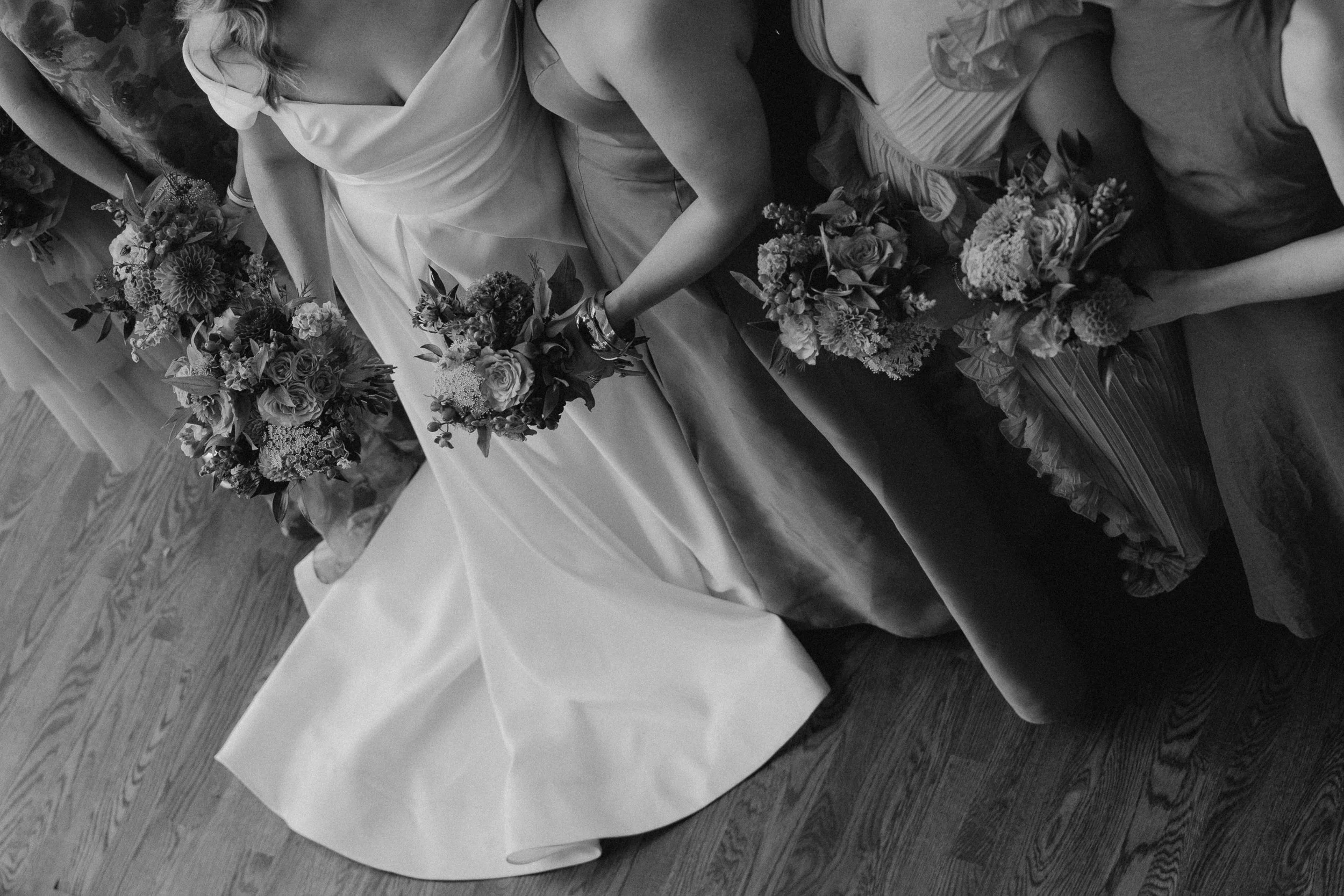 Black and white photo of women in dresses holding bouquets of flowers, sitting on a wooden floor, participating in a wedding or special event.