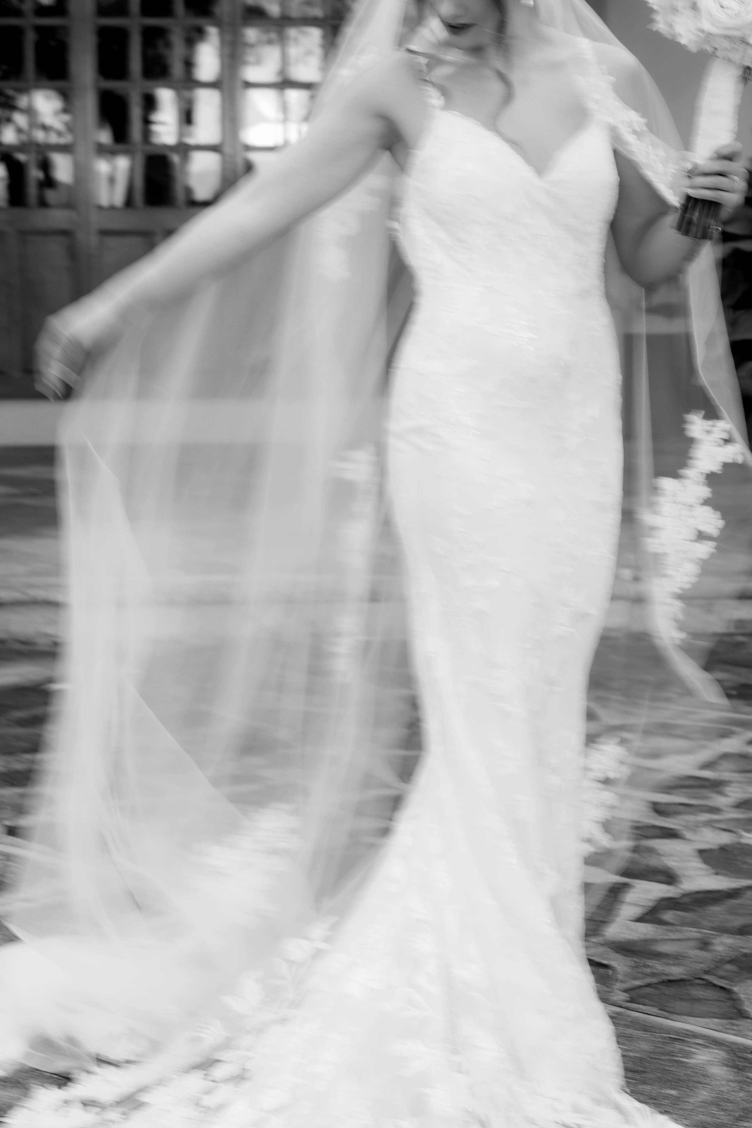 A bride in a white wedding dress with a long train and veil, holding a bouquet, walking on a patterned floor in a decorated venue.