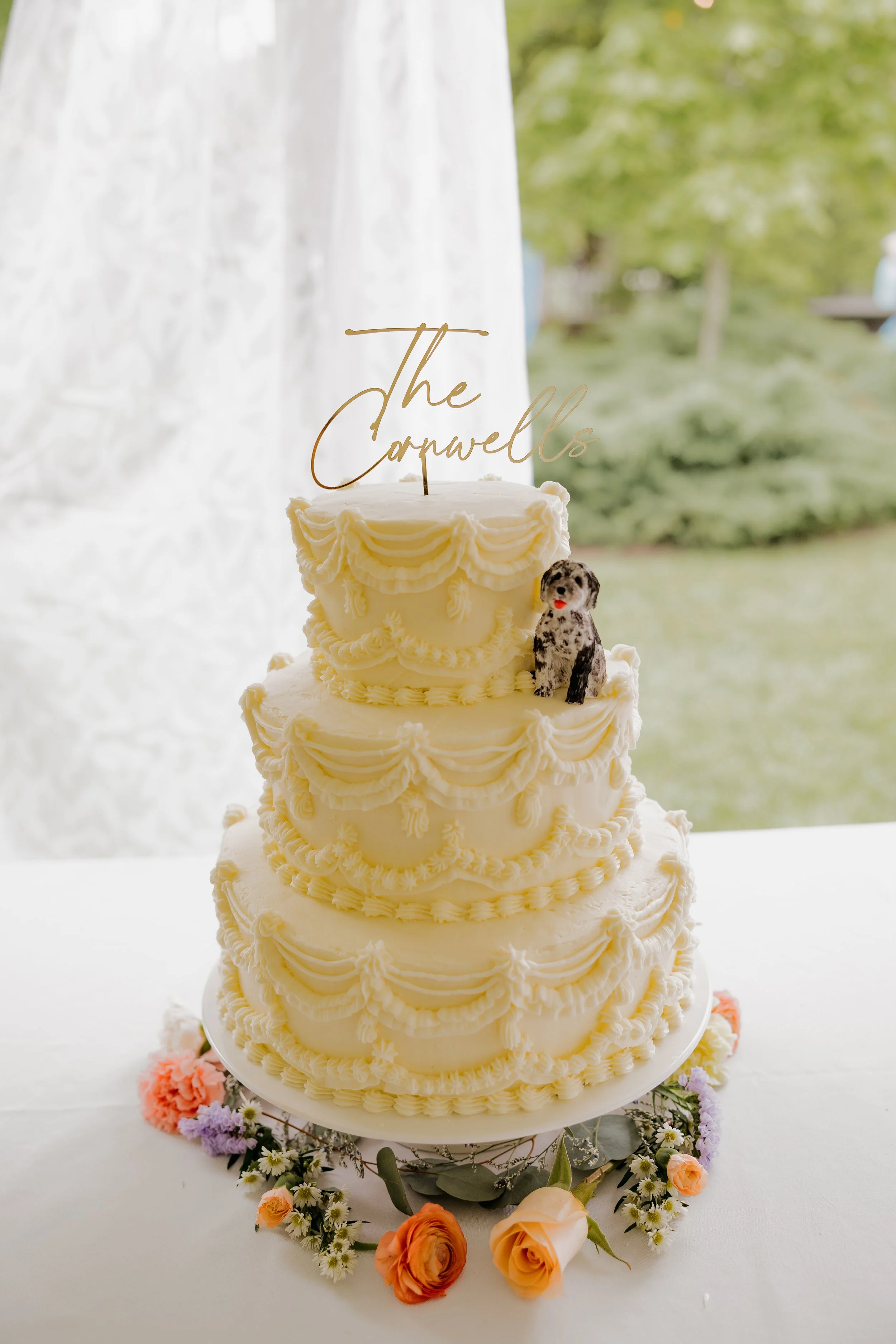 Three-tiered wedding cake with white icing and decorative piped borders, adorned with a topper reading 'The Conwells', a small dog figurine, and surrounded by flowers.