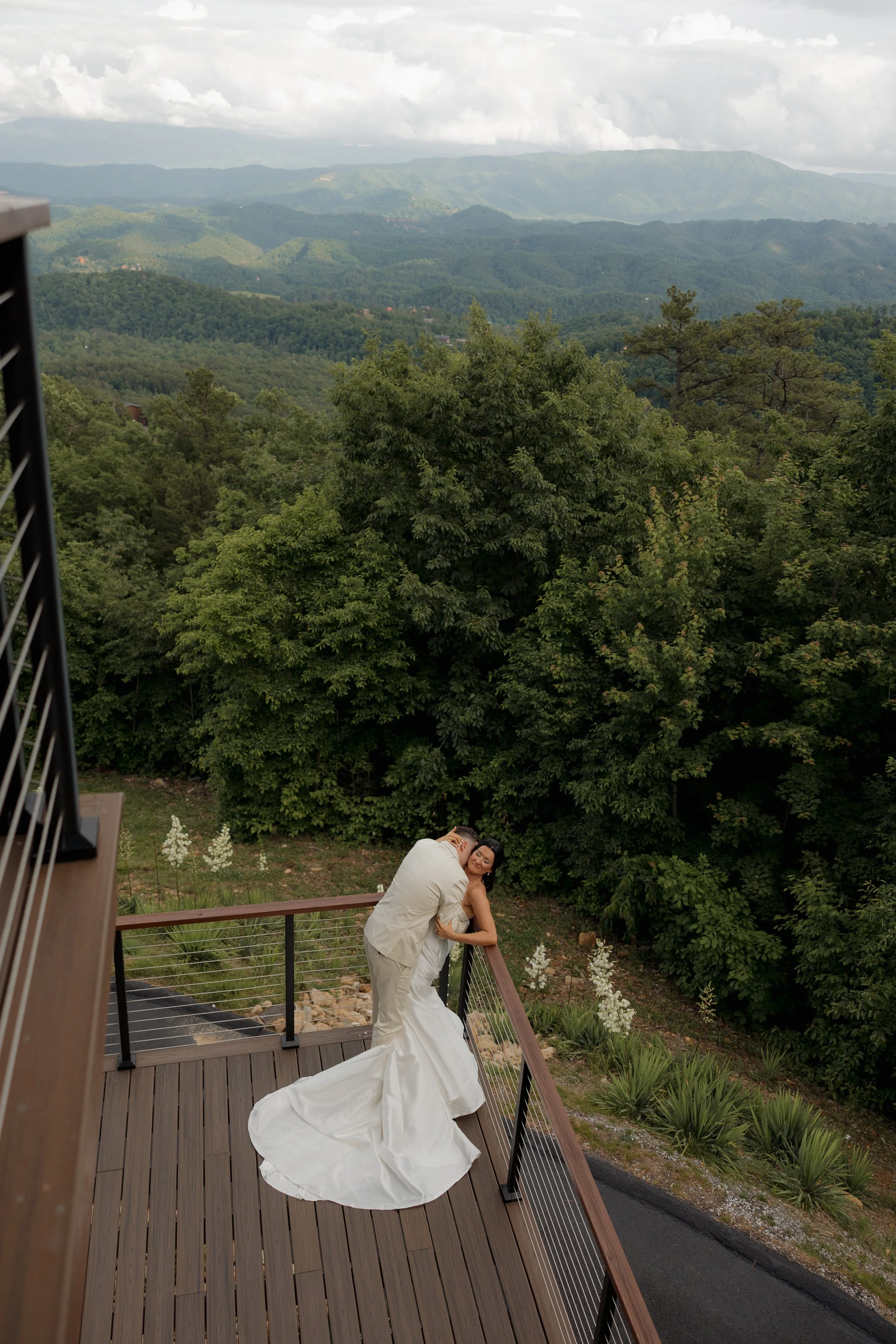 A bride and groom embrace on a wooden balcony overlooking lush green trees and mountain ranges in the distance, under a cloudy sky.