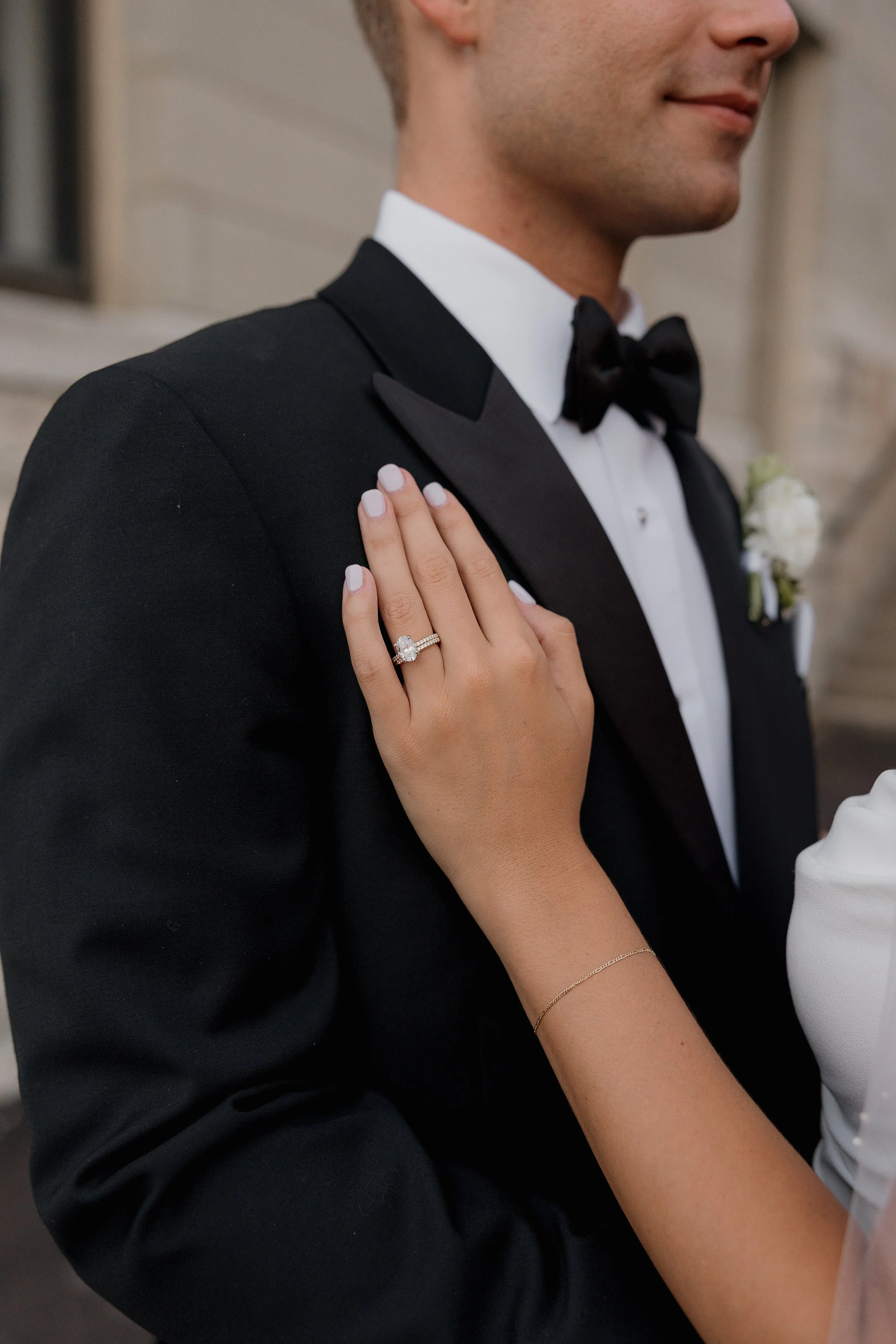 Close-up of a woman’s hand with an engagement ring on her ring finger resting on a man’s shoulder during a wedding or formal event.