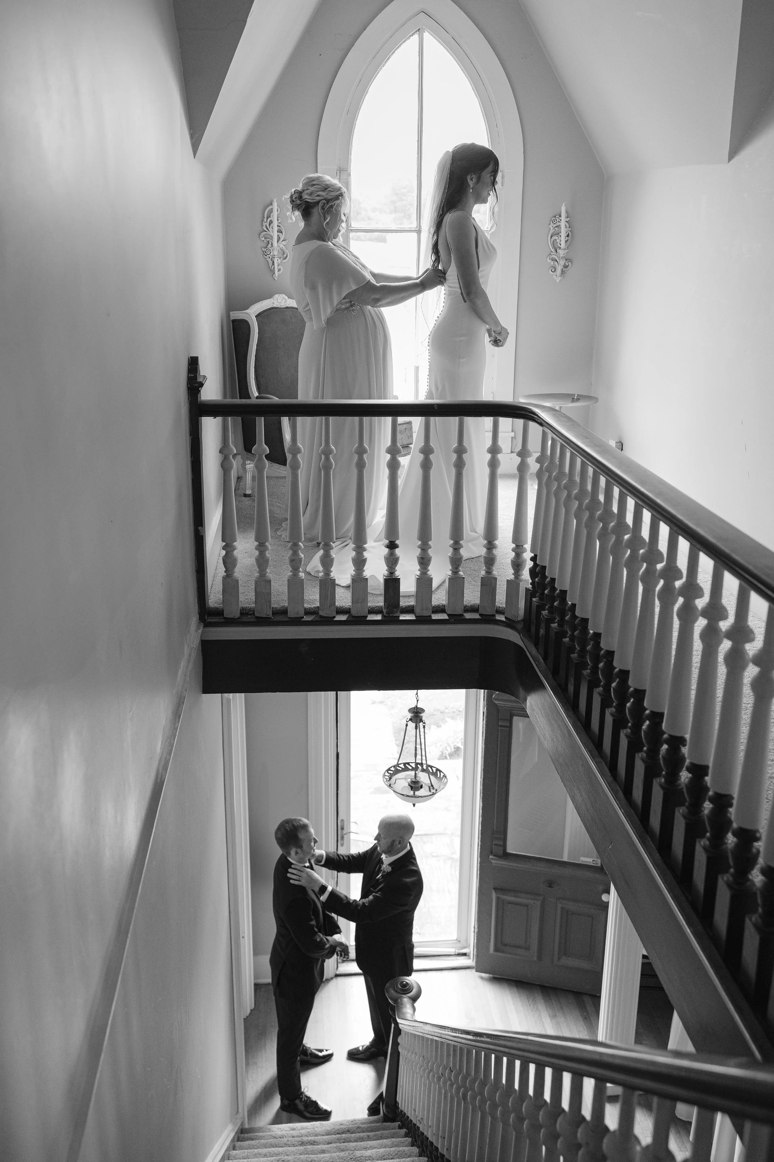 Black and white photo of a woman in a wedding dress standing on an upstairs balcony, with an older woman helping her with the dress. Below, two men in suits embrace at the bottom of the stairs by the front door.