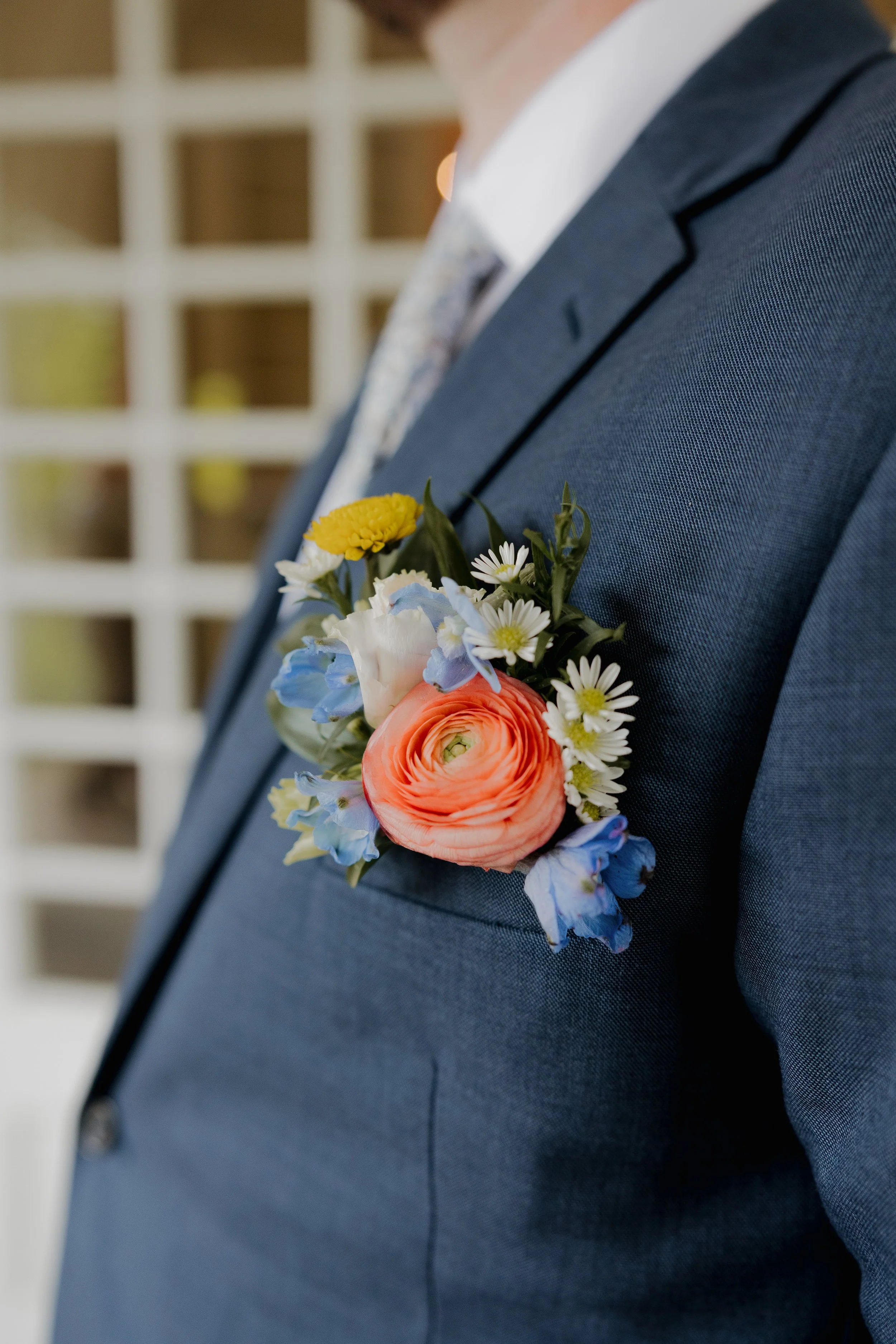 Close-up of a man wearing a navy suit with a colorful boutonniere pinned to his lapel, consisting of peach, white, blue, and yellow flowers.