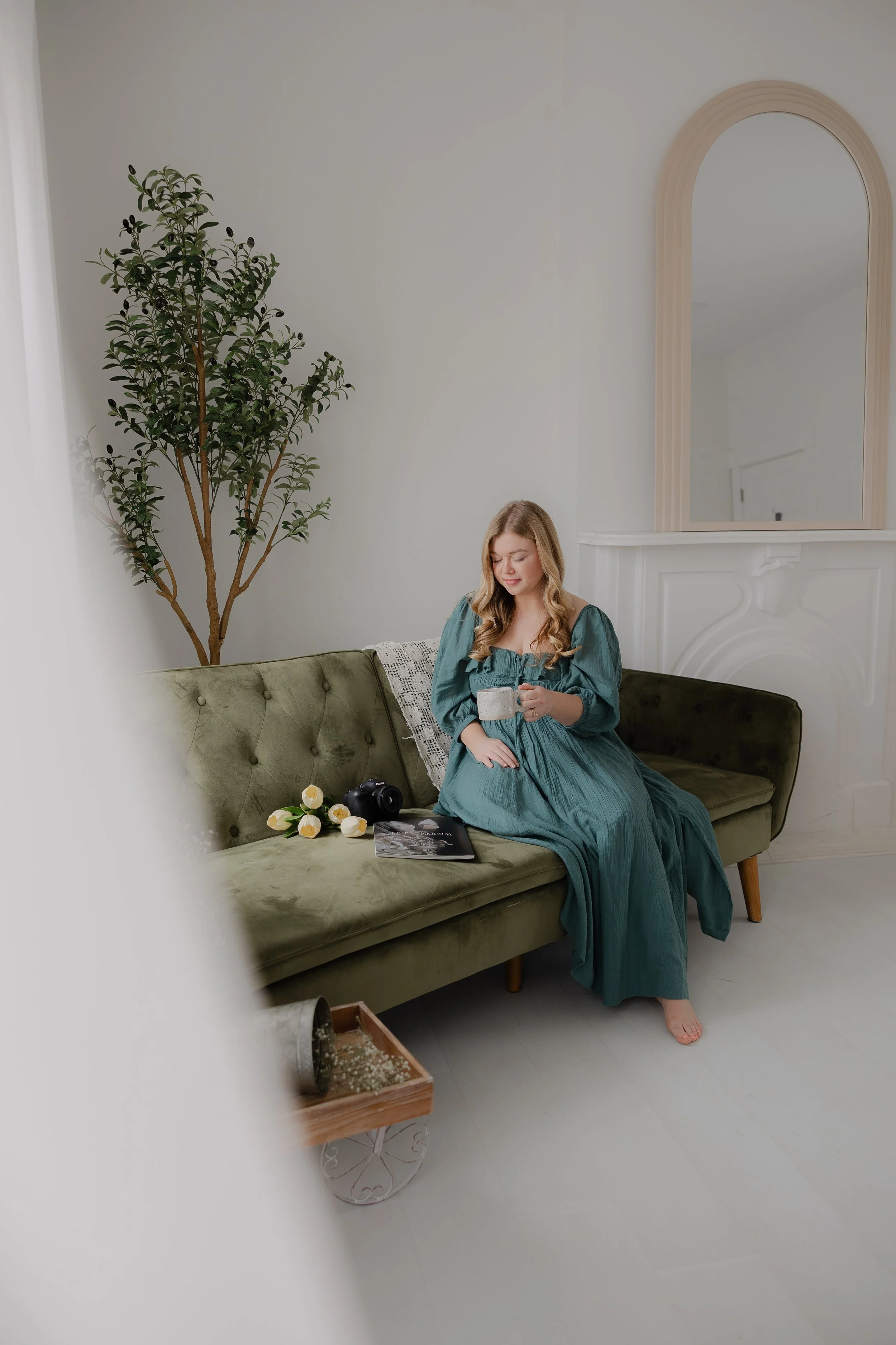 A woman with long blonde hair in a teal dress sitting on a green velvet couch, holding a mug, in a minimalist room with a large mirror and a potted plant in the background. Items on the couch include a camera, flowers, and a magazine.