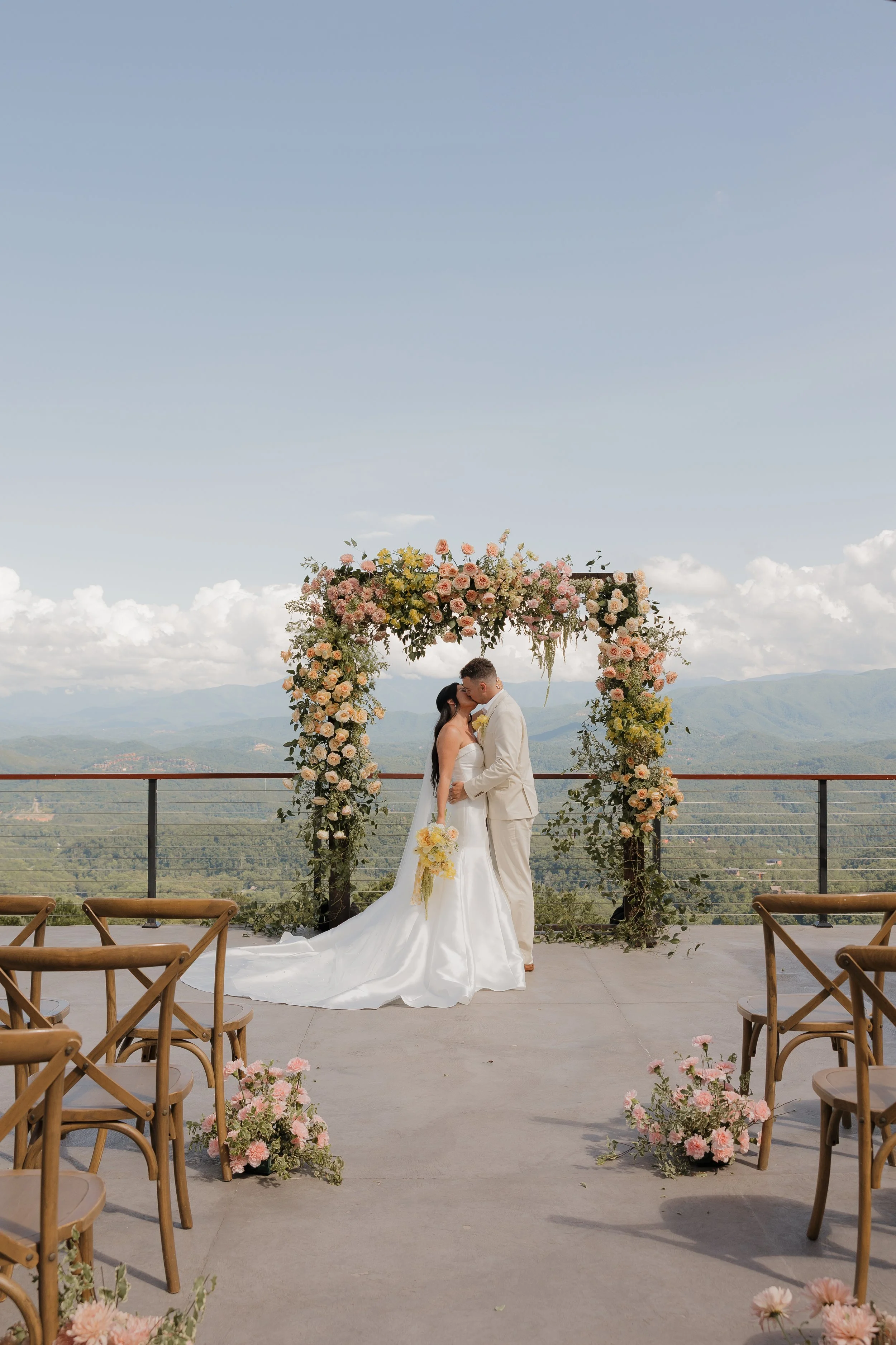 A bride and groom kissing under a floral wedding arch on an outdoor terrace with mountain views.
