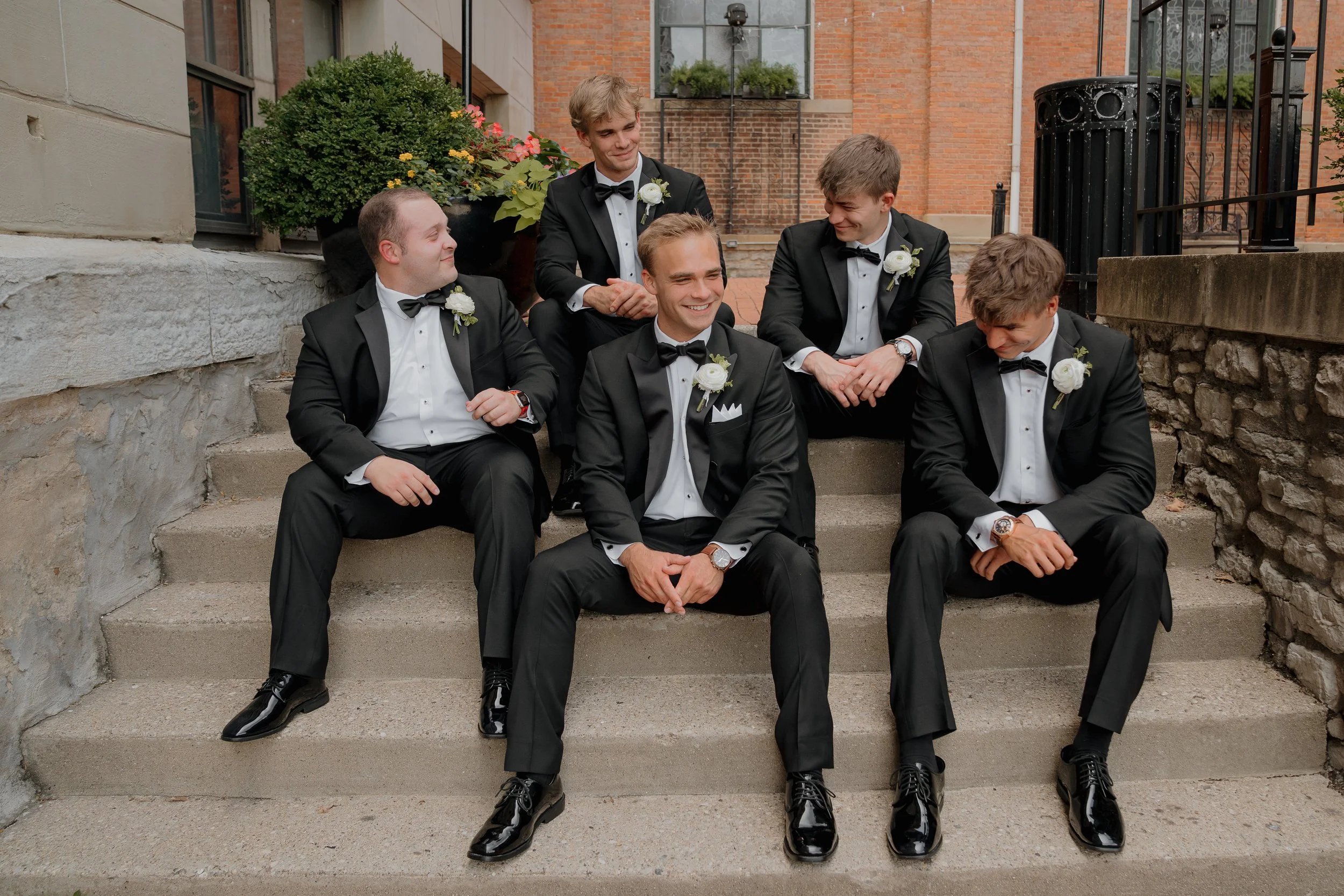 Group of six men dressed in black tuxedos with white shirts and black bow ties, sitting on outdoor steps, smiling and looking at each other, with a brick building and potted plants in the background.