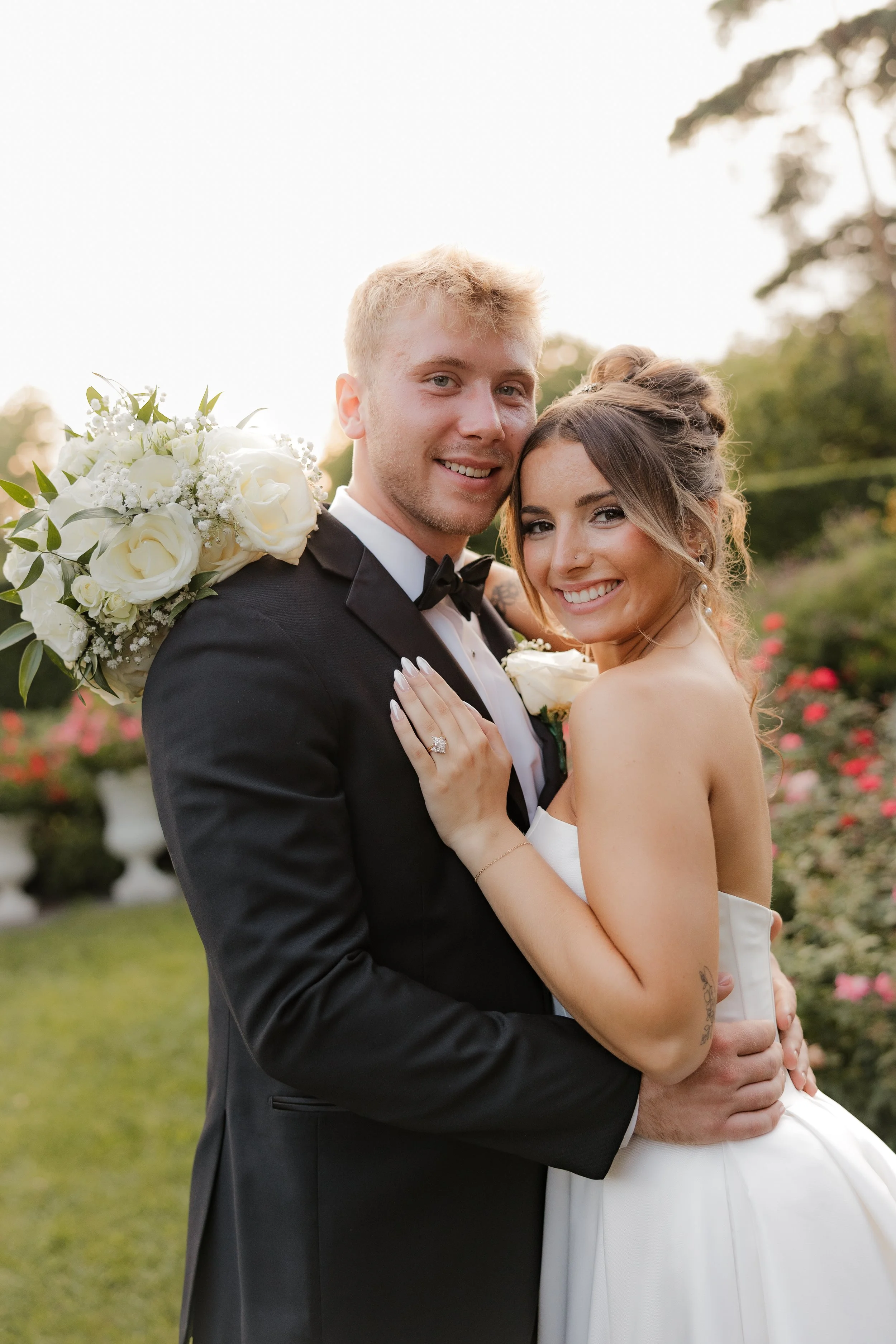 Wedding couple smiling, the groom in a black tuxedo and the bride in a strapless white wedding dress, embracing outdoors with flowers and greenery in the background.