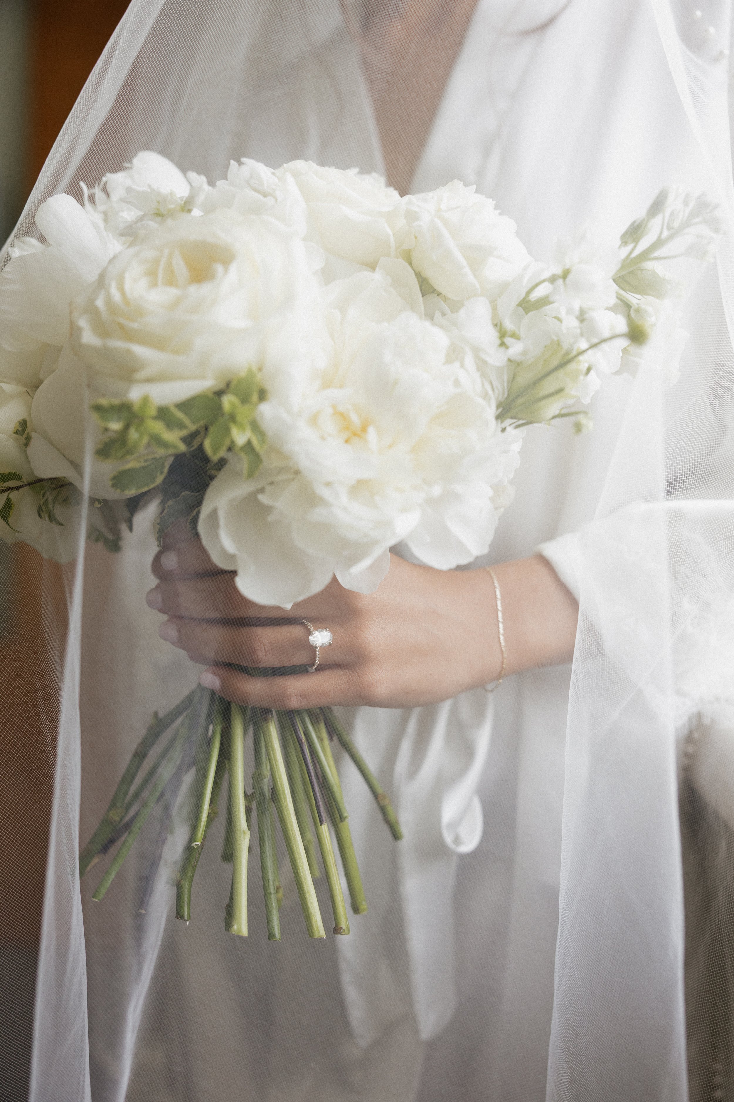 A bride holding a bouquet of white roses and peonies, wearing a wedding dress and a veil.