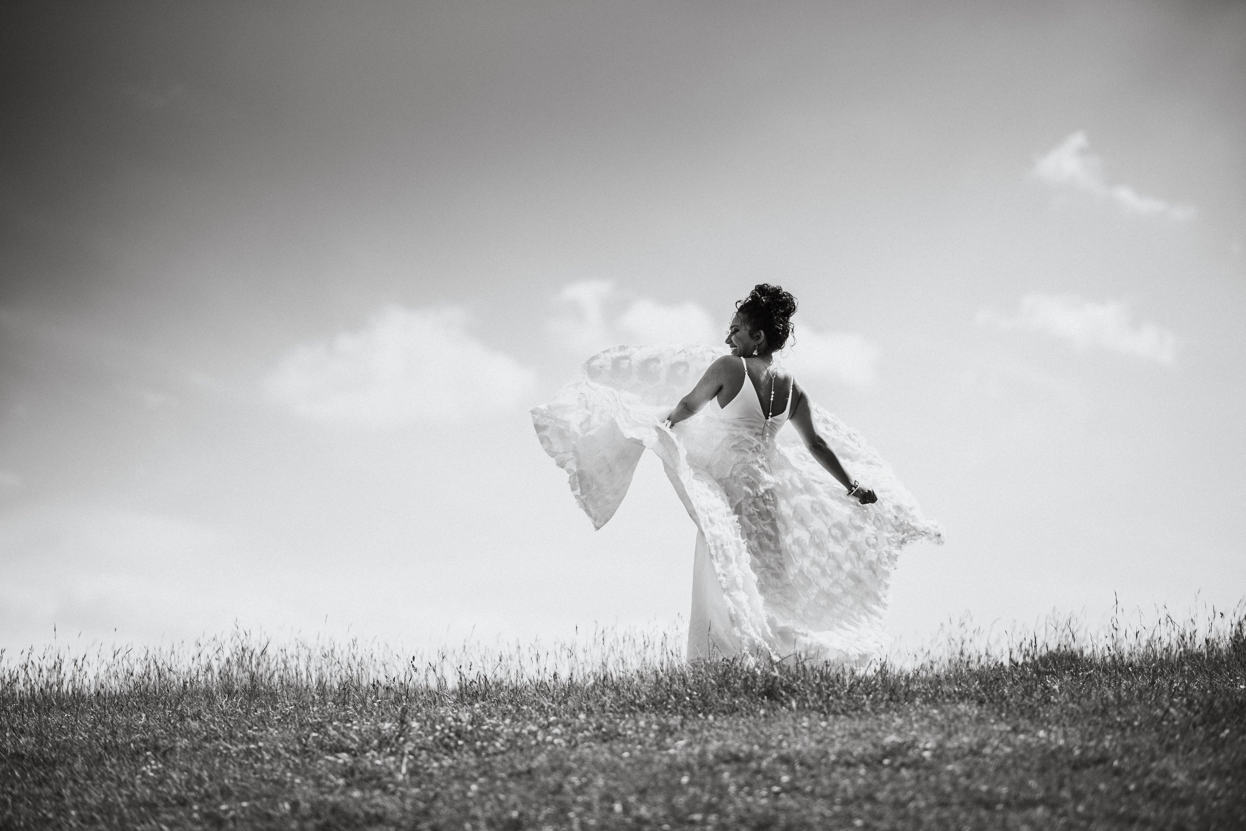 A woman in a flowing dress standing on a grassy hill under a partly cloudy sky, facing away from the camera with her head turned to the side.
