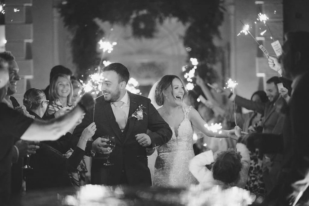 A black and white photo of a wedding celebration with the bride and groom walking through guests holding sparklers.