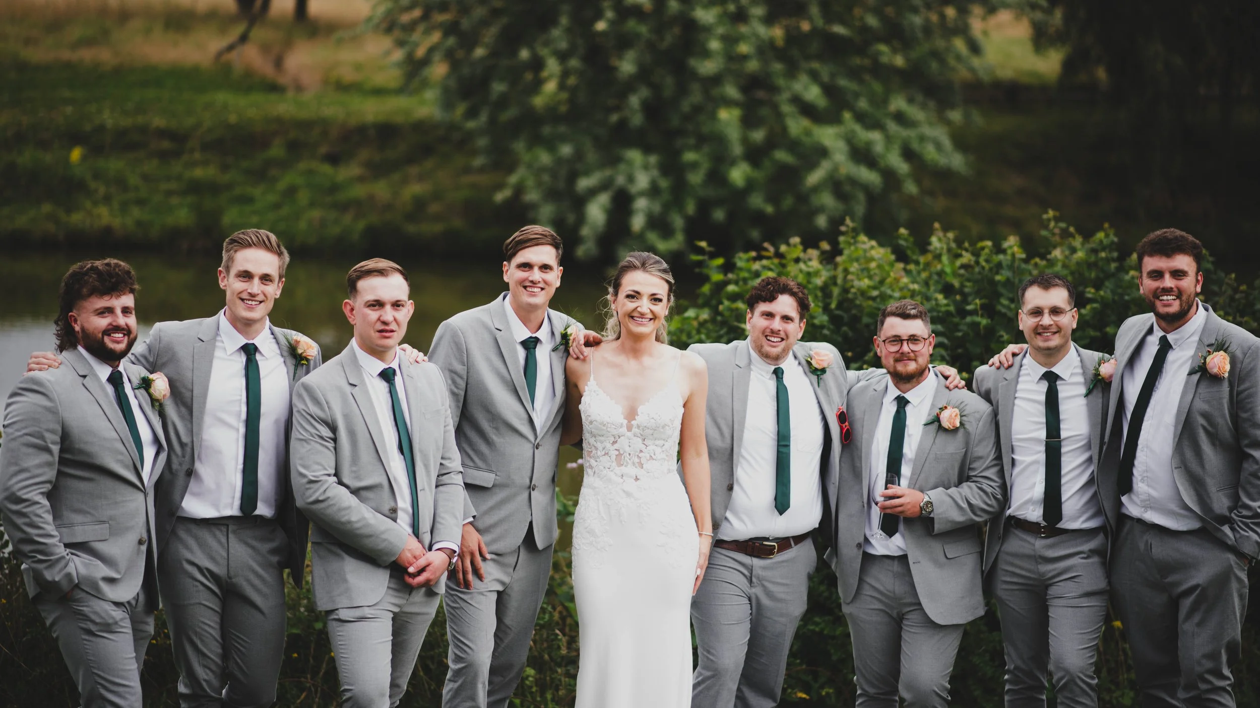 A bride and eight groomsmen standing outdoors on a grassy area with trees and a body of water in the background. The bride is in a white lace wedding gown, and the groomsmen are in gray suits with white shirts, green ties, and pink boutonnieres.