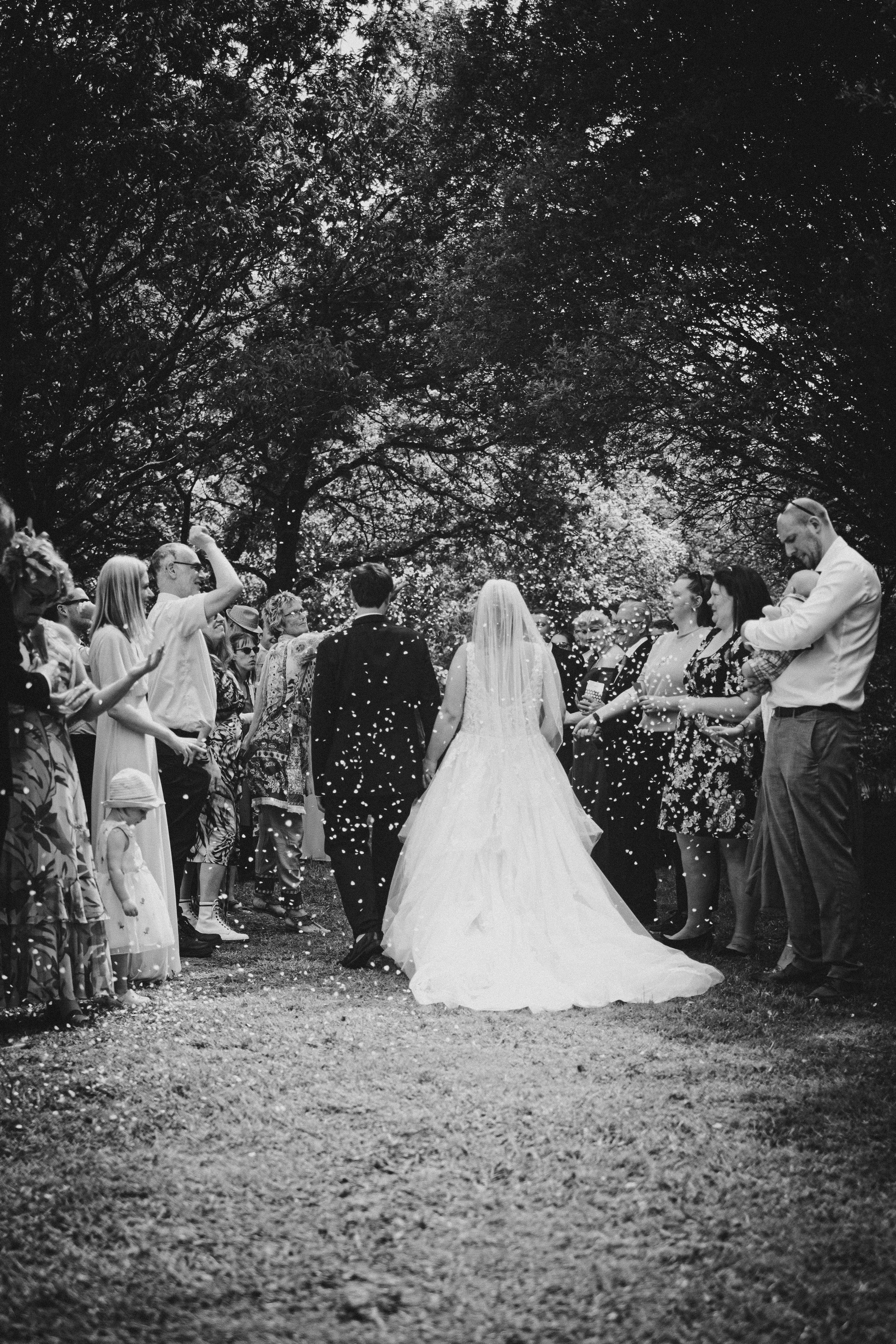 Black and white photo of a bride and groom walking away from a crowd of friends and family during an outdoor wedding, surrounded by trees.