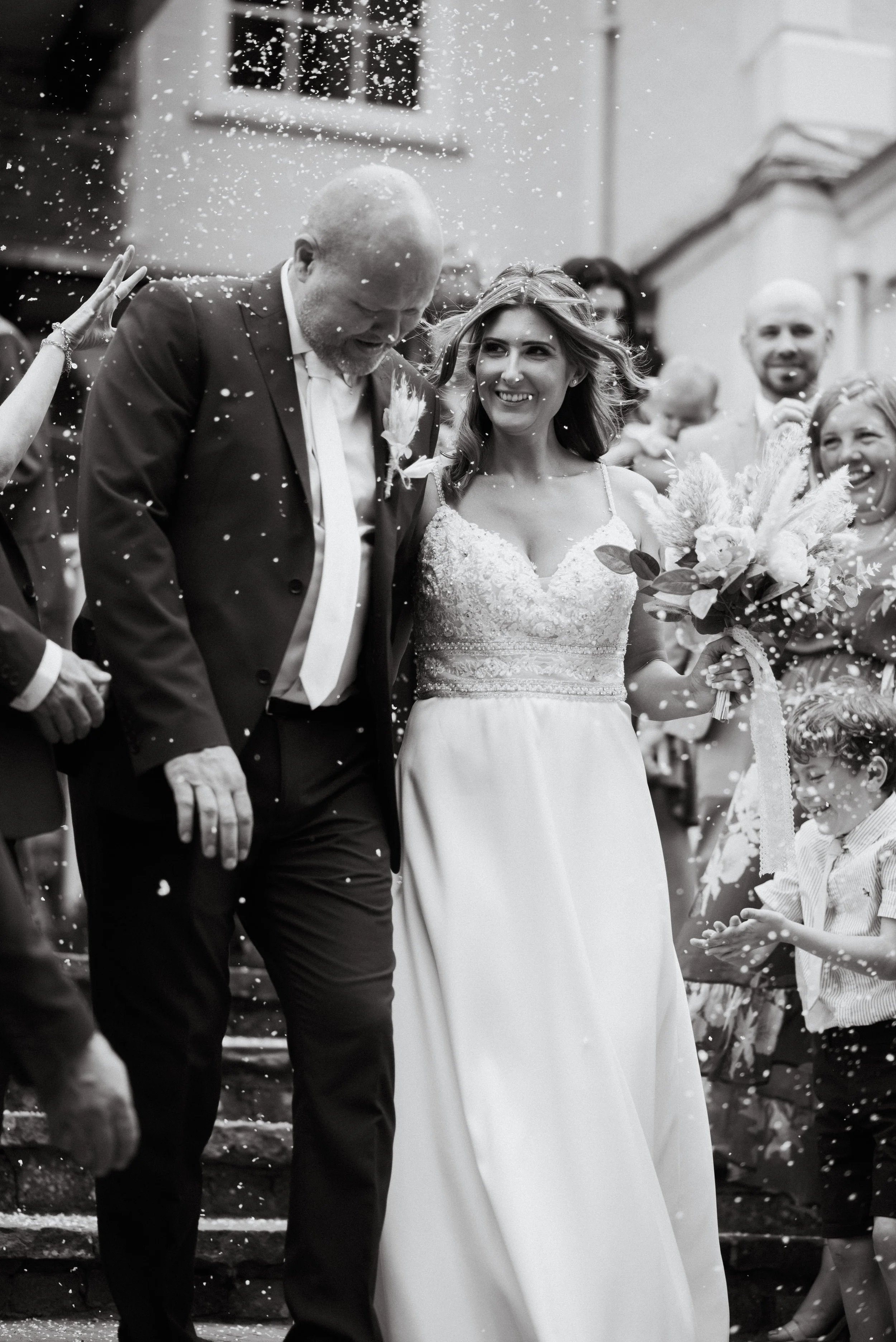 A black and white photo of a wedding celebration outside a building. The bride and groom are walking together, smiling, surrounded by friends and family. The bride is holding a bouquet, and guests are throwing rice or confetti.