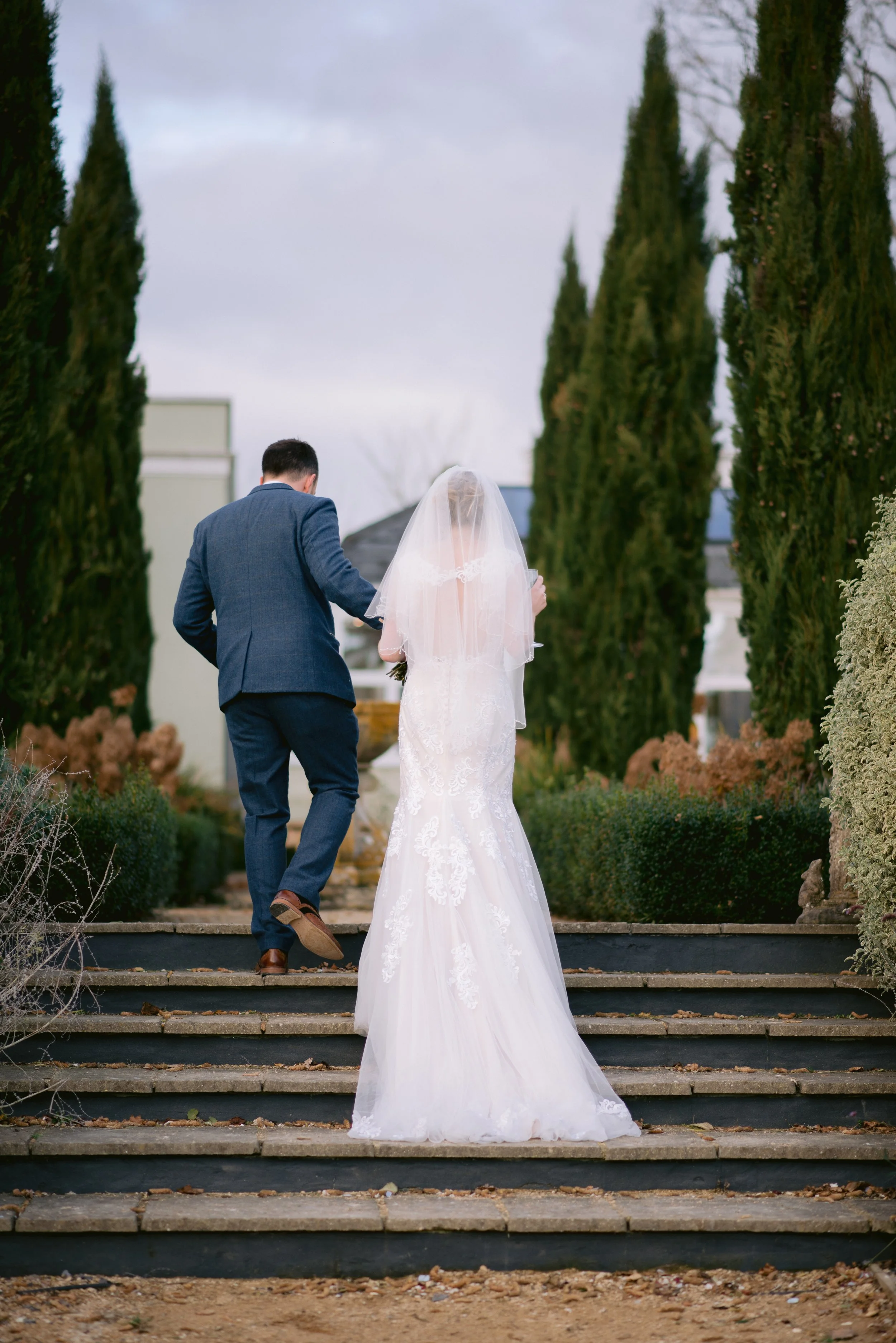 A bride and groom walking up steps outside, surrounded by trees and bushes, on their wedding day.